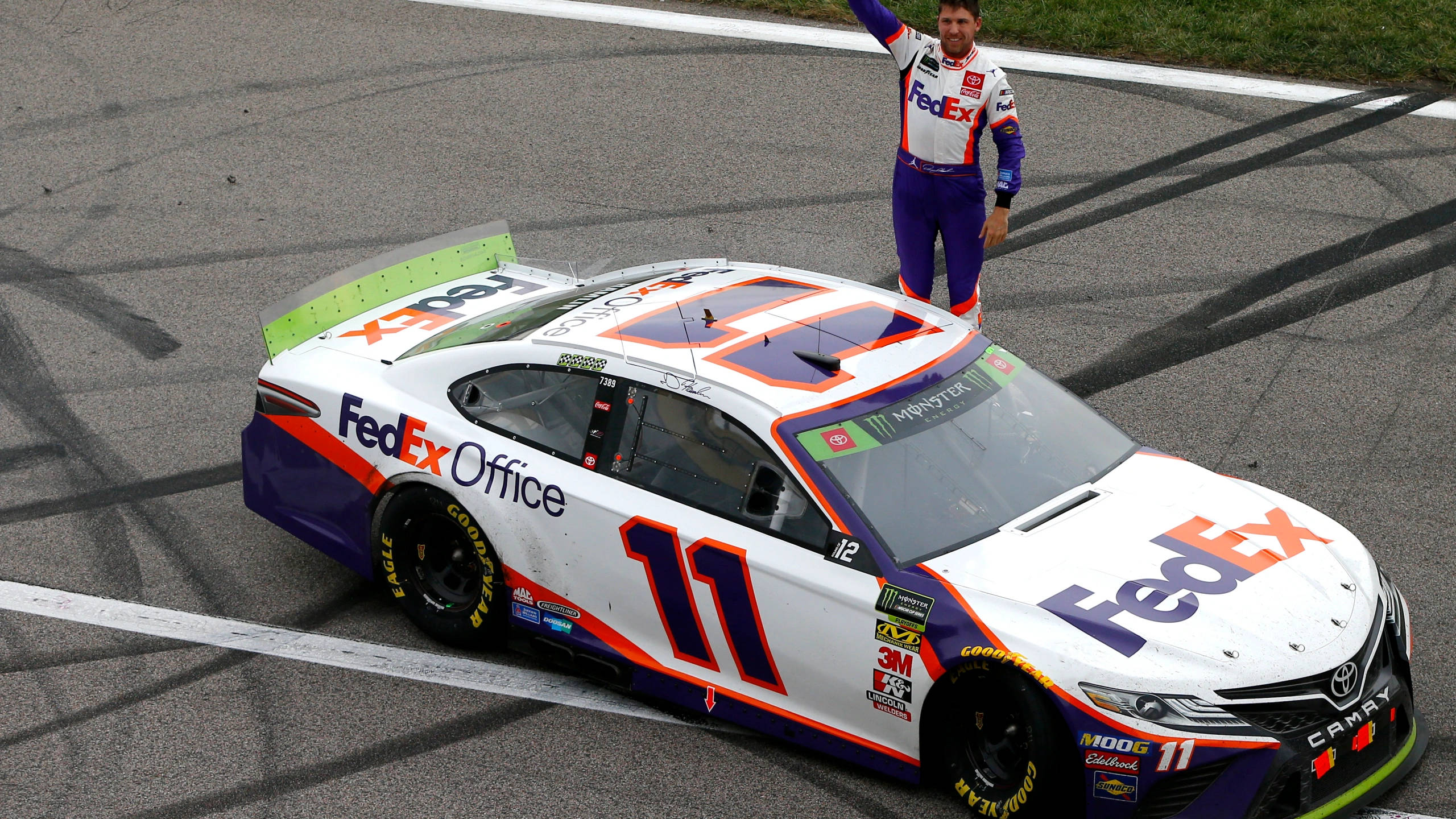 Denny Hamlin 桌布 壁紙 — 一名身穿FedEx制服的NASCAR車手，在他白色、紫色和橙色的#11 FedEx Office賽車頂上慶祝，背景是賽道。