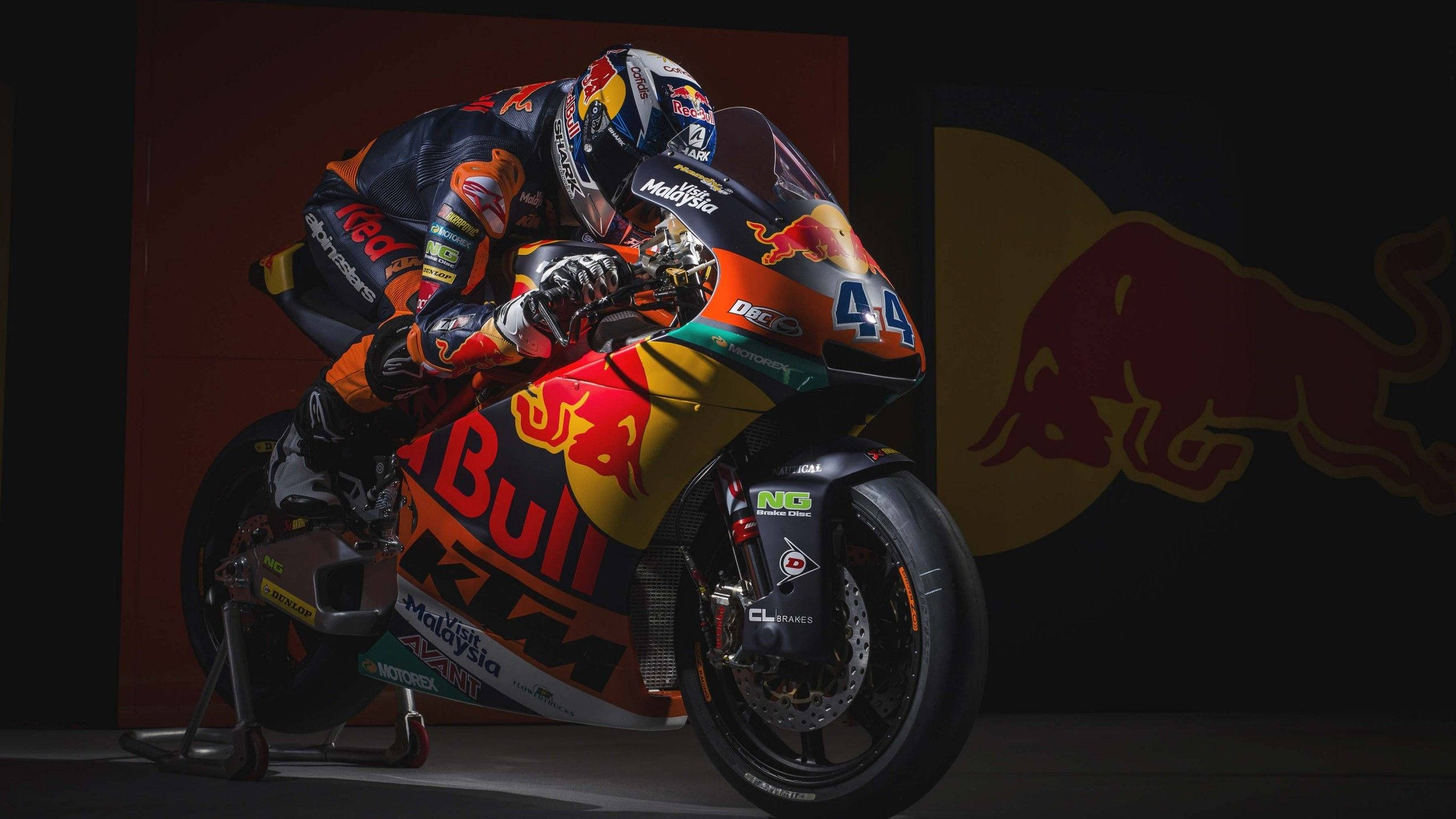 Red Bull 桌布 壁紙 — 一名身穿Red Bull KTM裝備的車手，在帶有大型Red Bull標誌的深色背景前擺出摩托車姿勢。