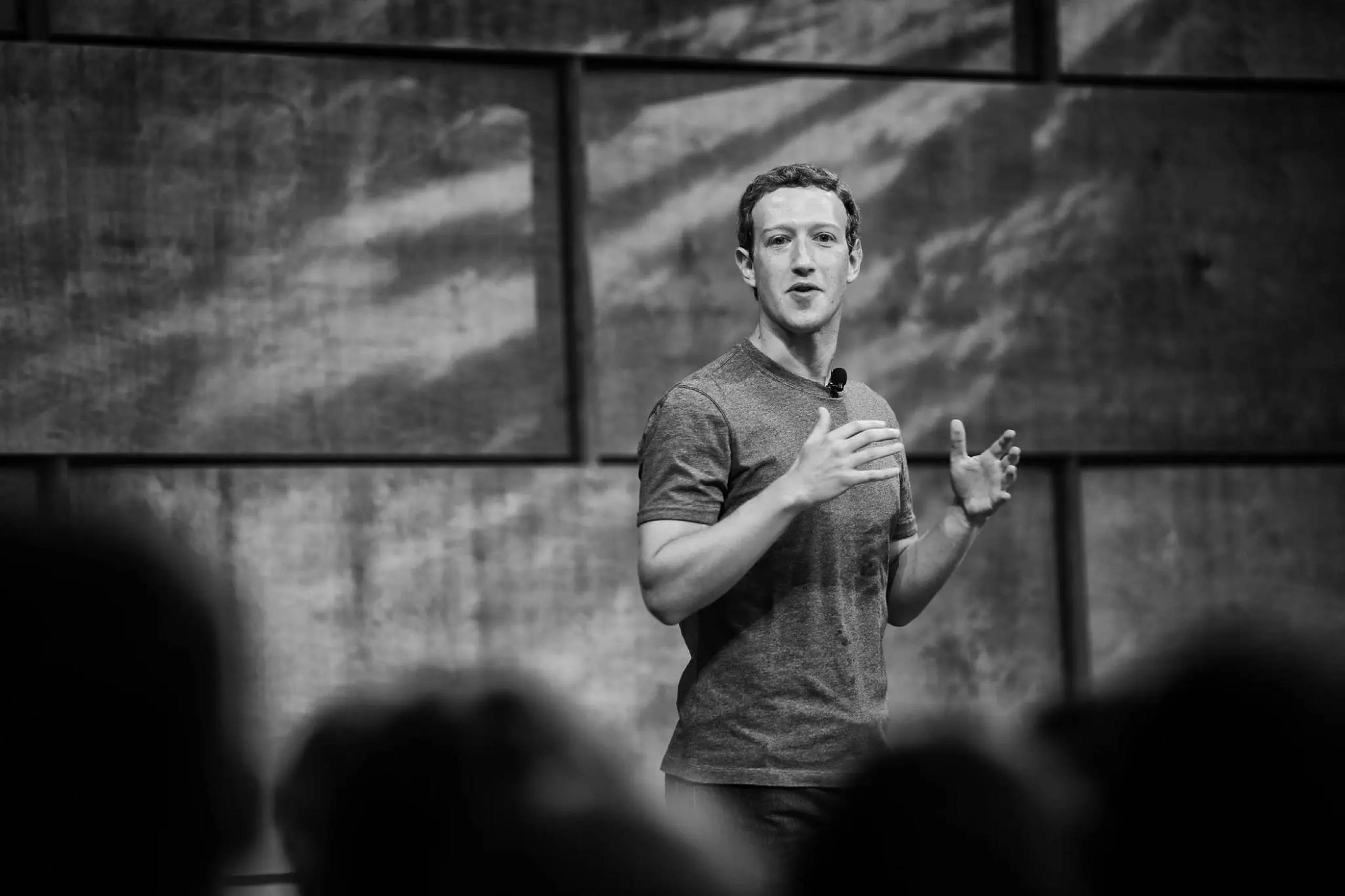 Zuckerberg 桌布 壁紙 — 一名身穿T恤的男子在舞台上熱情演講，模糊的觀眾剪影以黑白呈現。