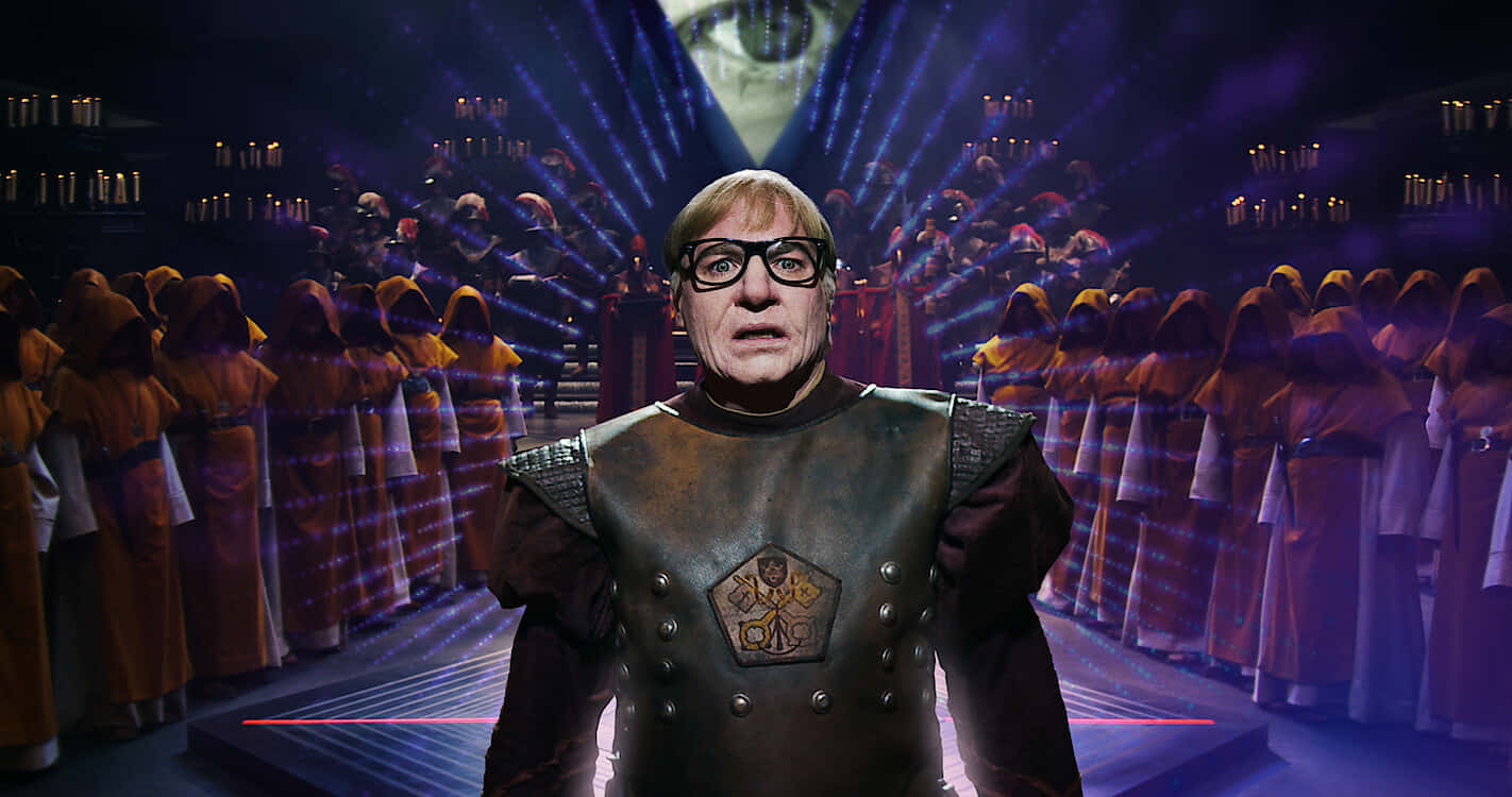 Mike Myers 桌布 壁紙 — 一名身穿中世紀盔甲的男人在紫色戲劇性聚光燈下，站在舞台中央，前面是一群穿著連帽長袍的合唱團。