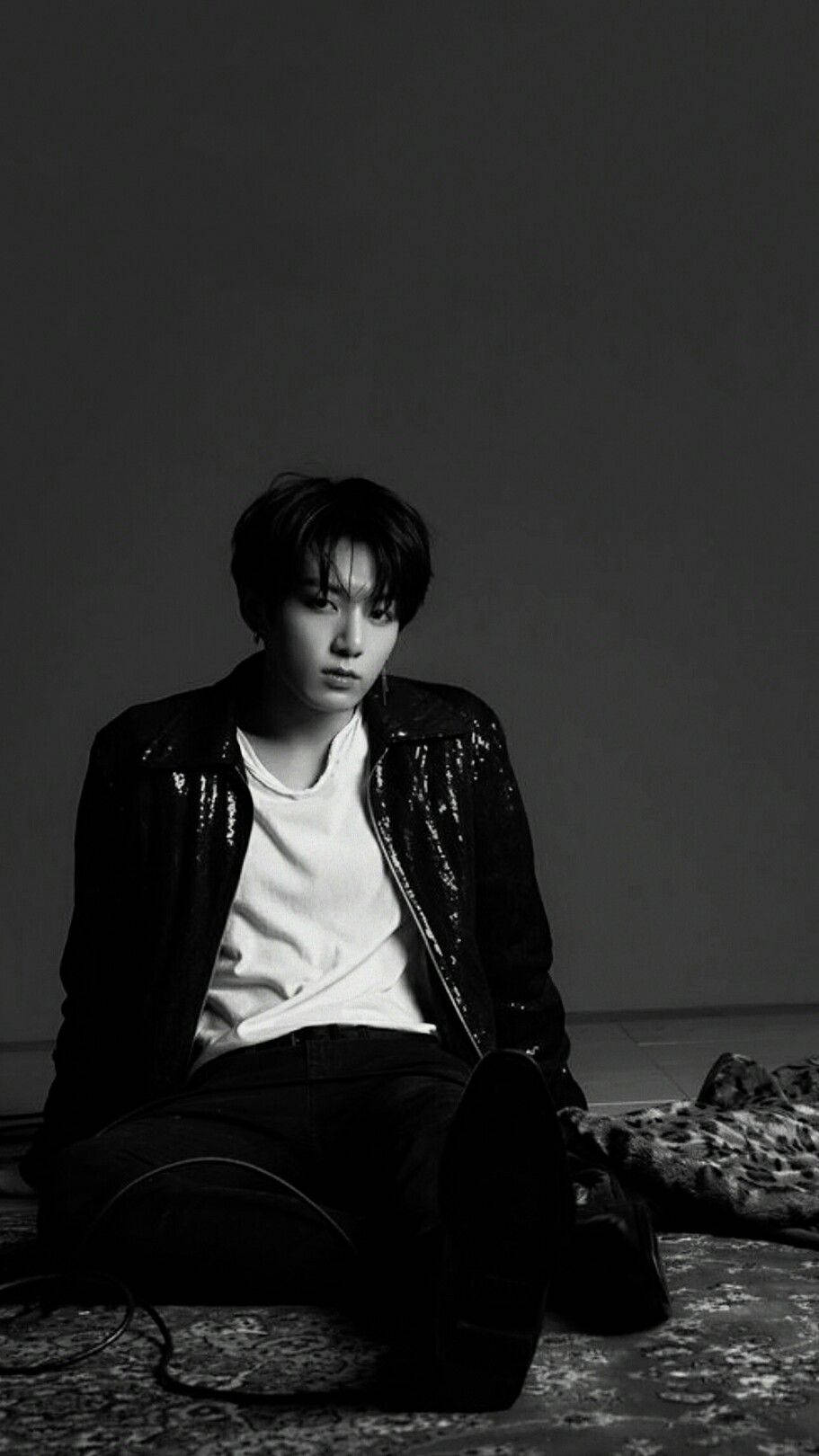 Jungkook 桌布 壁紙 — 一名身穿亮片外套的年輕男子坐在圖案地毯上，靠著素色牆壁，呈現出陰鬱的黑白肖像。