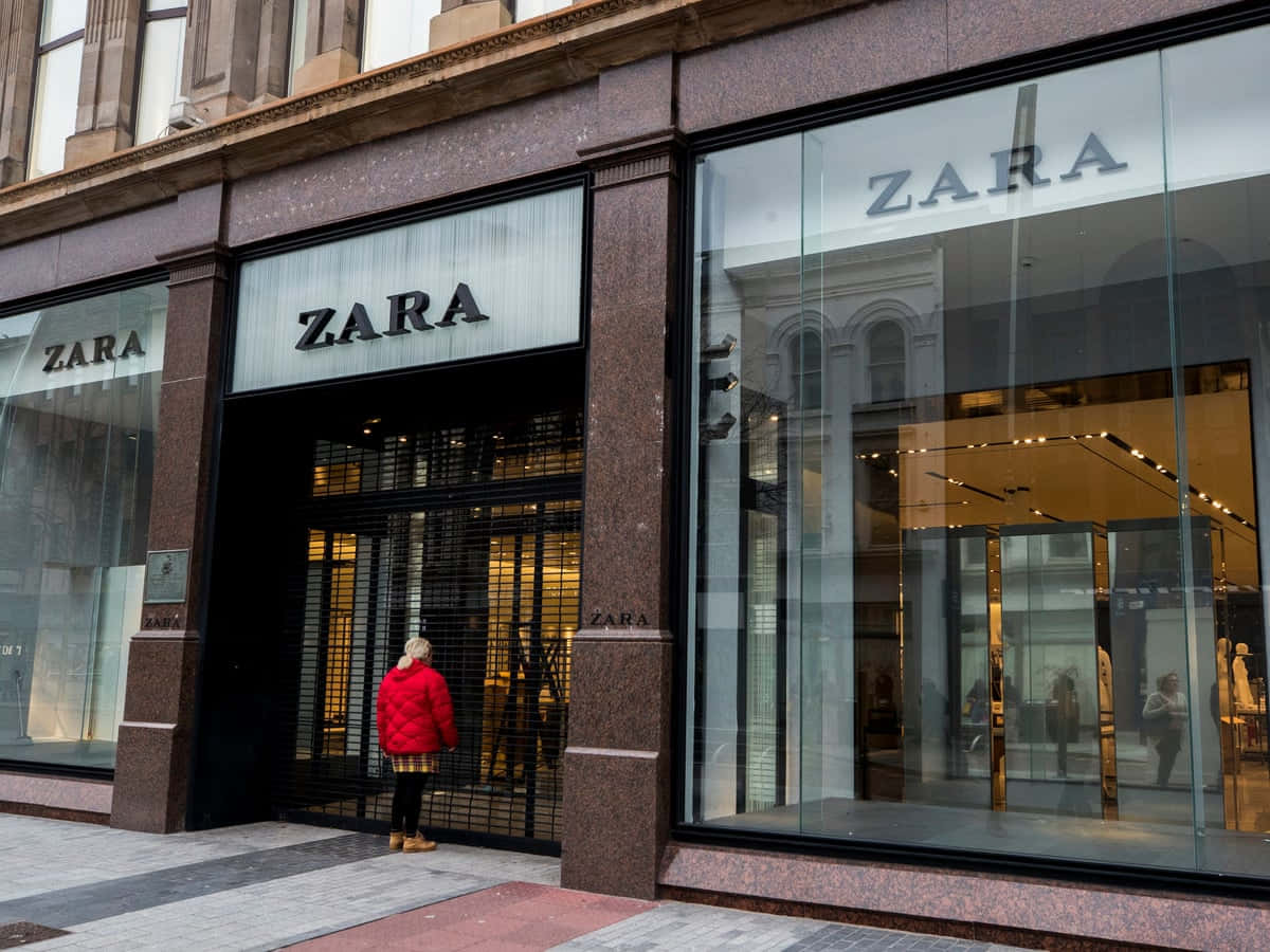 Zara 桌布 壁紙 — 一名身穿亮紅色外套的人站在Zara店鋪的玻璃入口前，店鋪有反光窗戶和石造外牆。