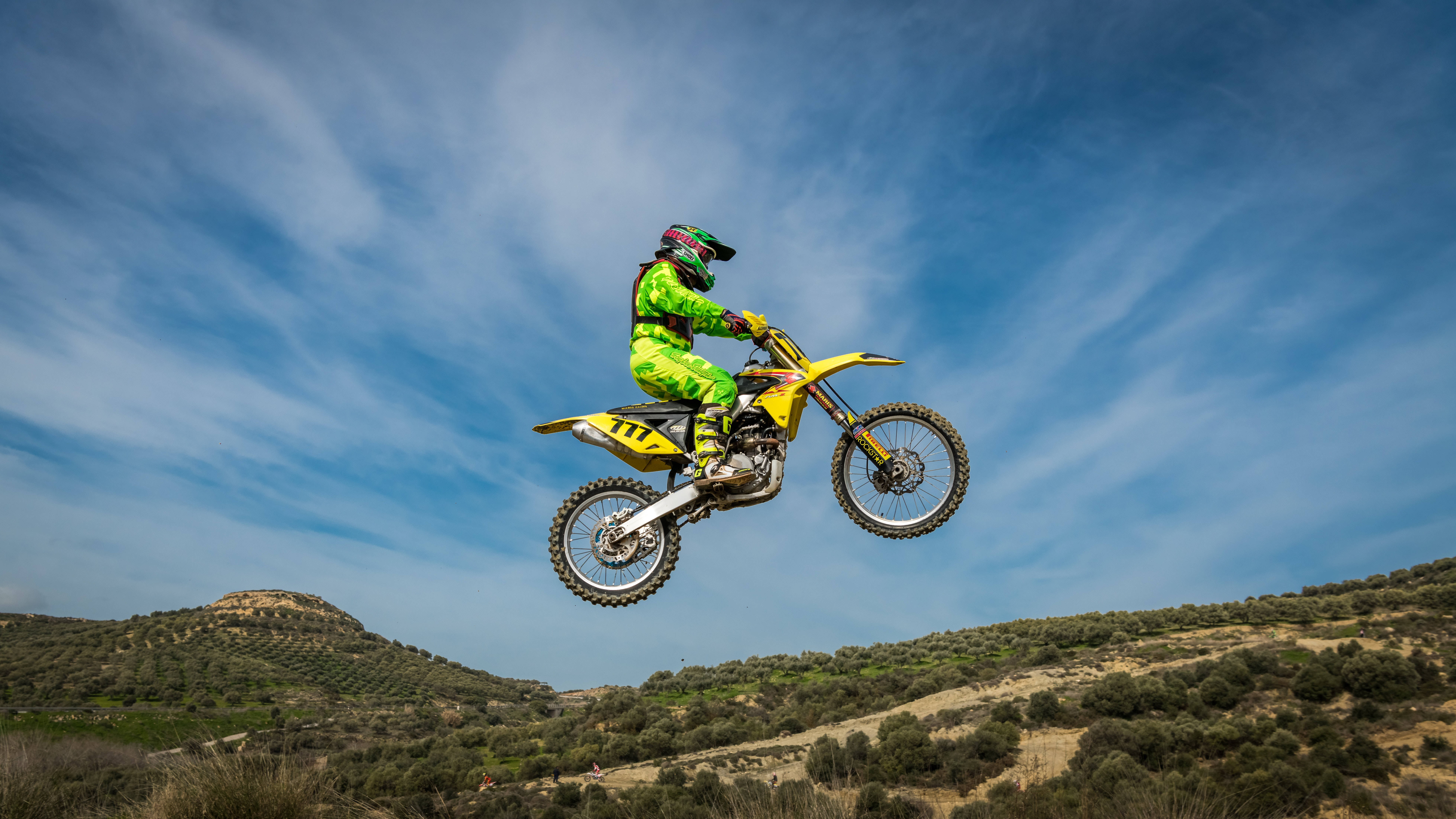 Motocross 桌布 壁紙 — 一名身穿亮綠色裝備的越野摩托車手，在廣闊的藍天白雲下，騎著黃色摩托車在空中飛翔。