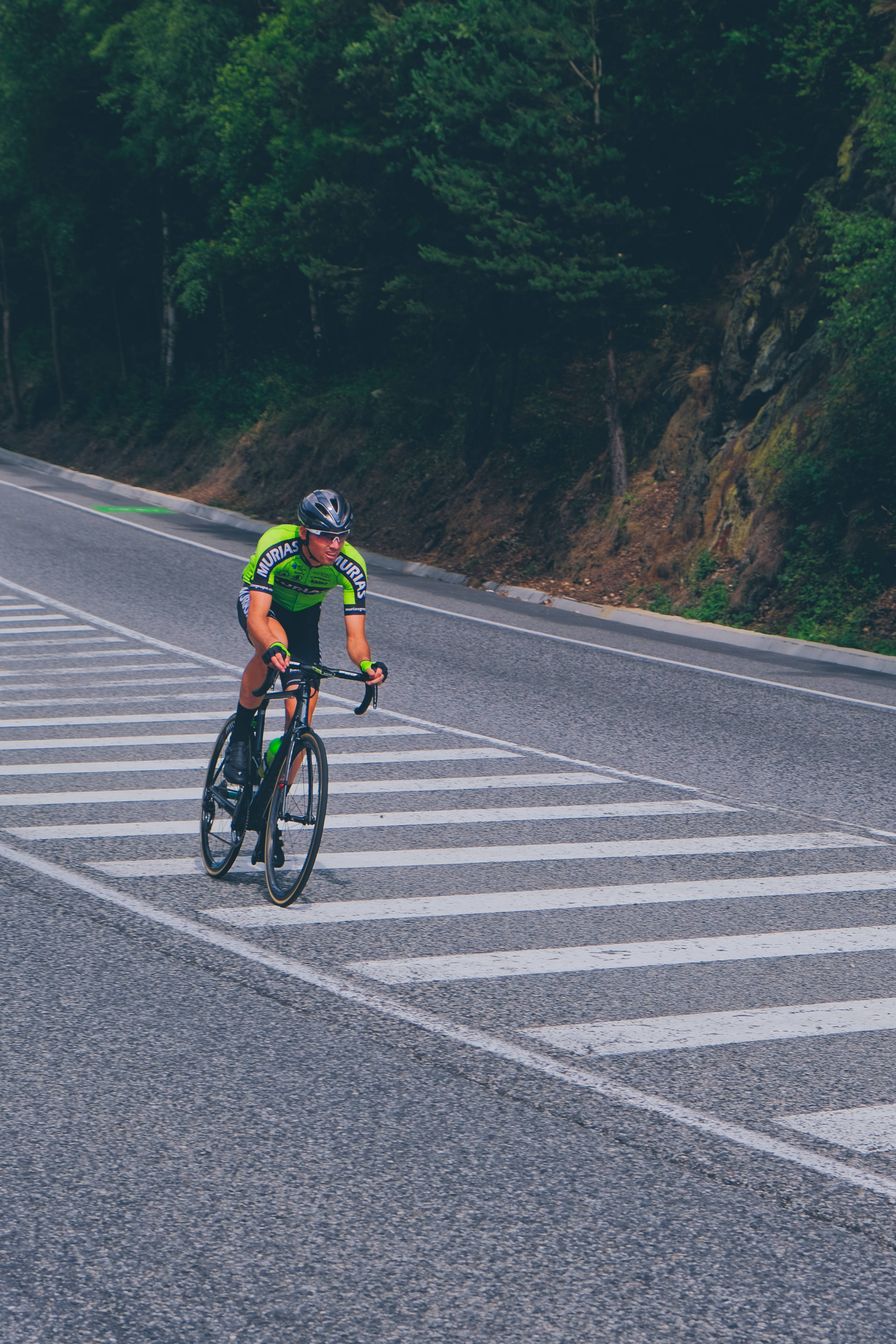 Bicycle 桌布 壁紙 — 一名身穿亮綠色運動衫的自行車手在蜿蜒的山路上，兩旁是茂密的森林，正用力踩著踏板穿過斑馬線。
