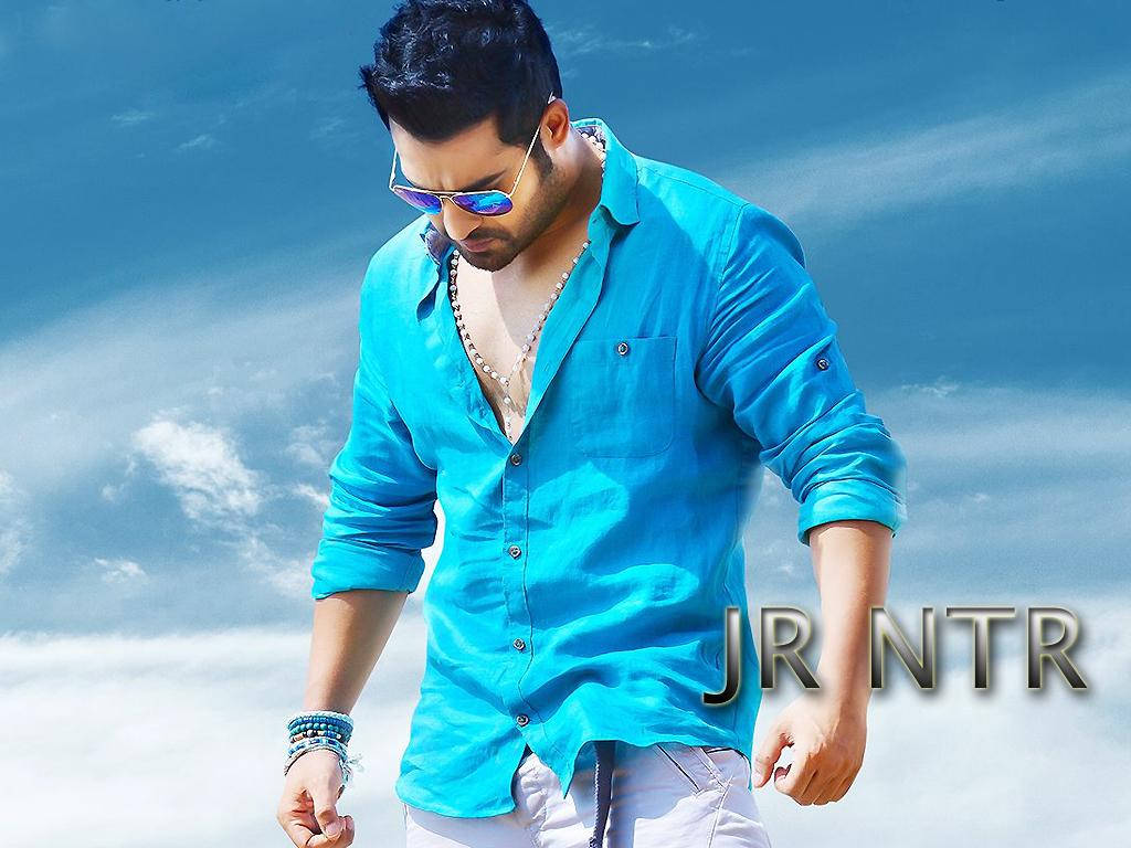 jr ntr 桌布 壁紙 — 一名身穿亮藍色襯衫和太陽眼鏡的男子，在帶有散雲的鮮豔藍天下自信地行走。