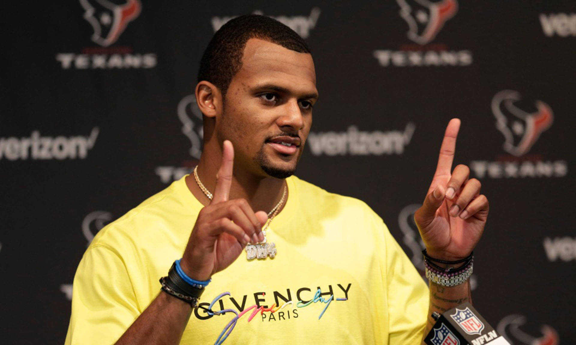 Deshaun Watson 桌布 壁紙 — 一名身穿亮黃色Givenchy T恤的男子在記者會上，以休士頓德州人隊為背景，舉起雙食指做手勢。