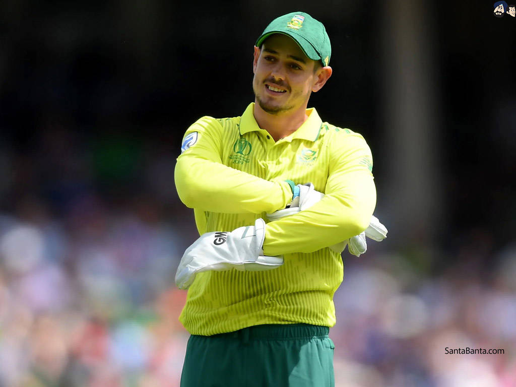 quinton de kock 桌布 壁紙 — 一名身穿亮黃色球衣和綠色帽子的板球運動員，雙臂交叉，自信地站在模糊的體育場人群前。