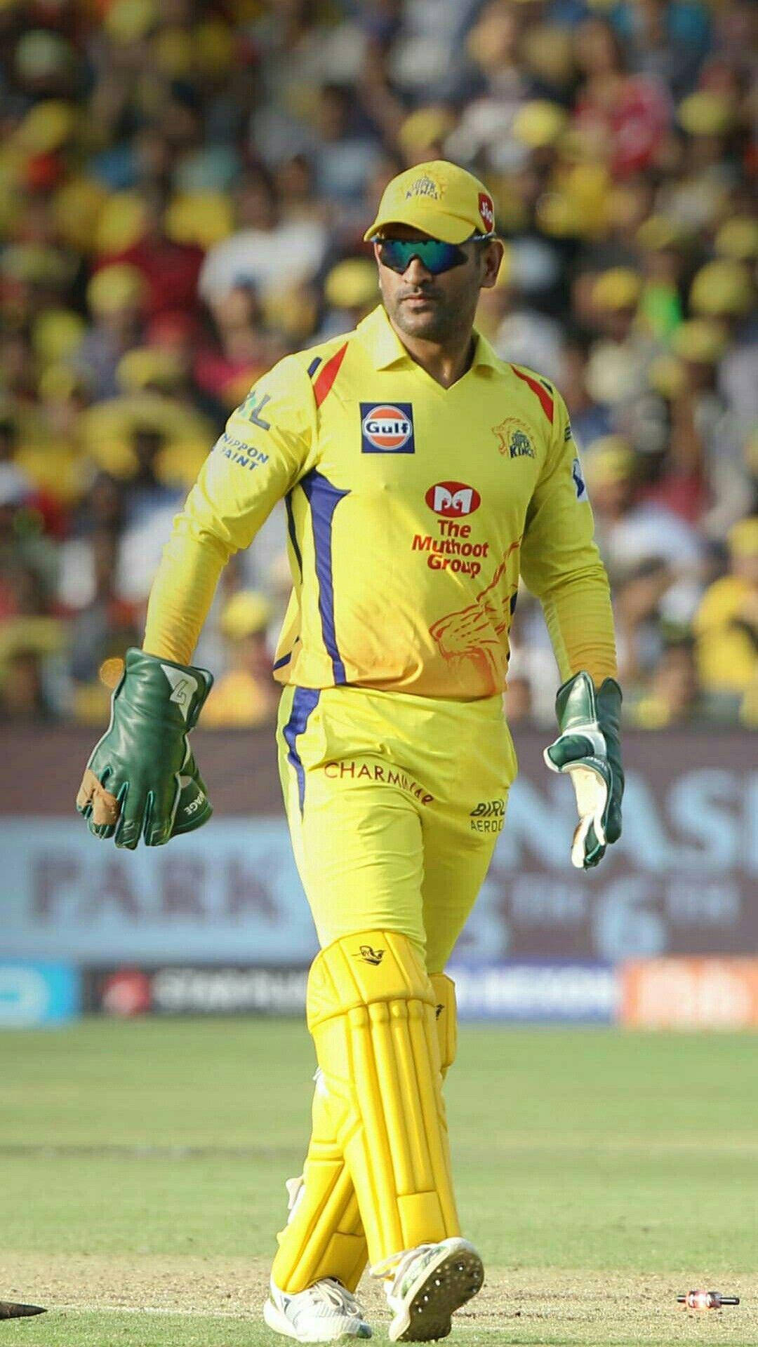 MS Dhoni 桌布 壁紙 — 一名身穿亮黃色球衣的板球運動員在綠色場地上行走，身後是戴著黃色帽子的模糊球迷人群。