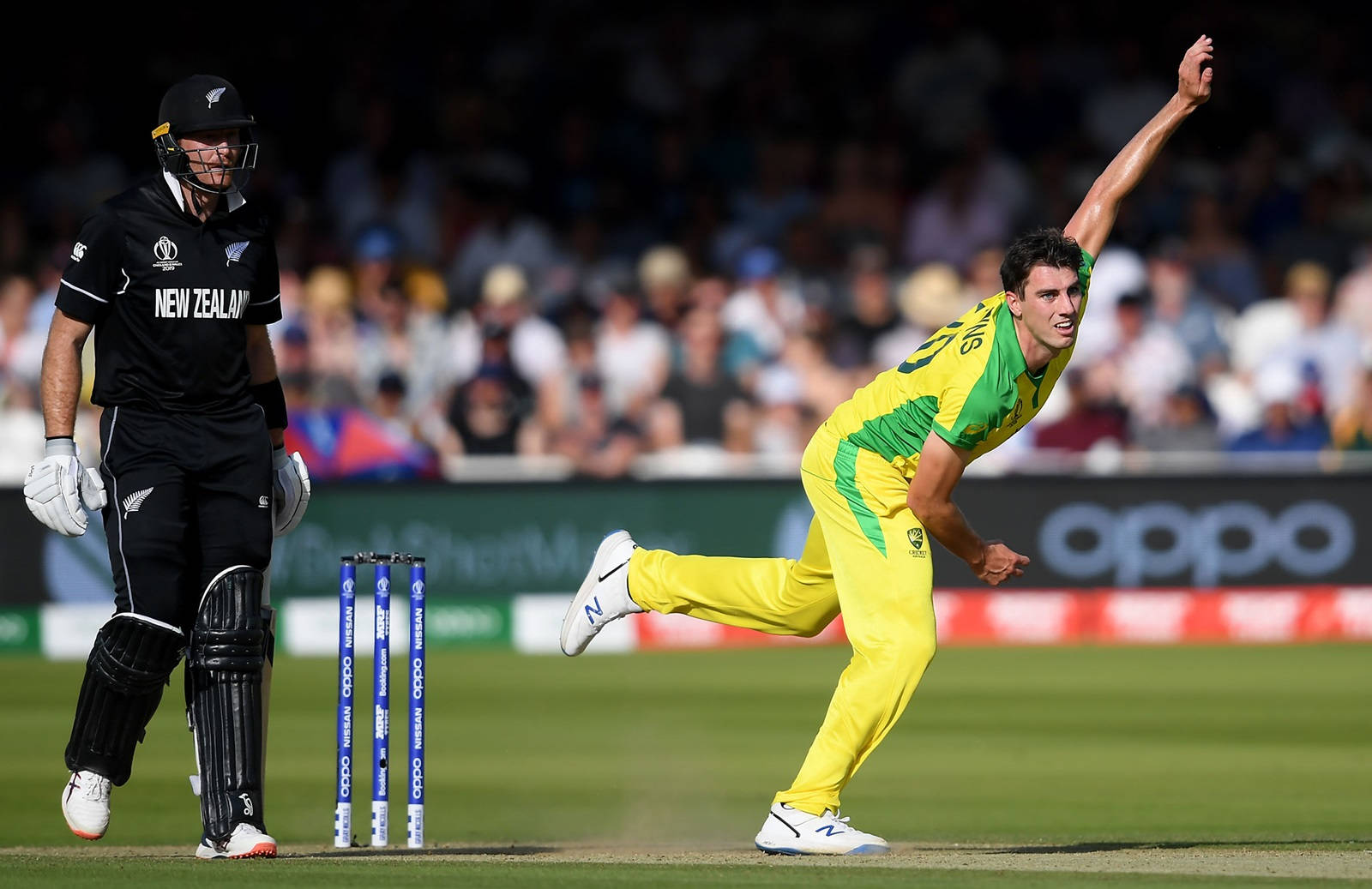 australia cricket 桌布 壁紙 — 一名身穿亮黃色的板球投球手正在投球，而一名身穿黑色的擊球手在陽光普照的綠色球場上的擊球線準備就緒。