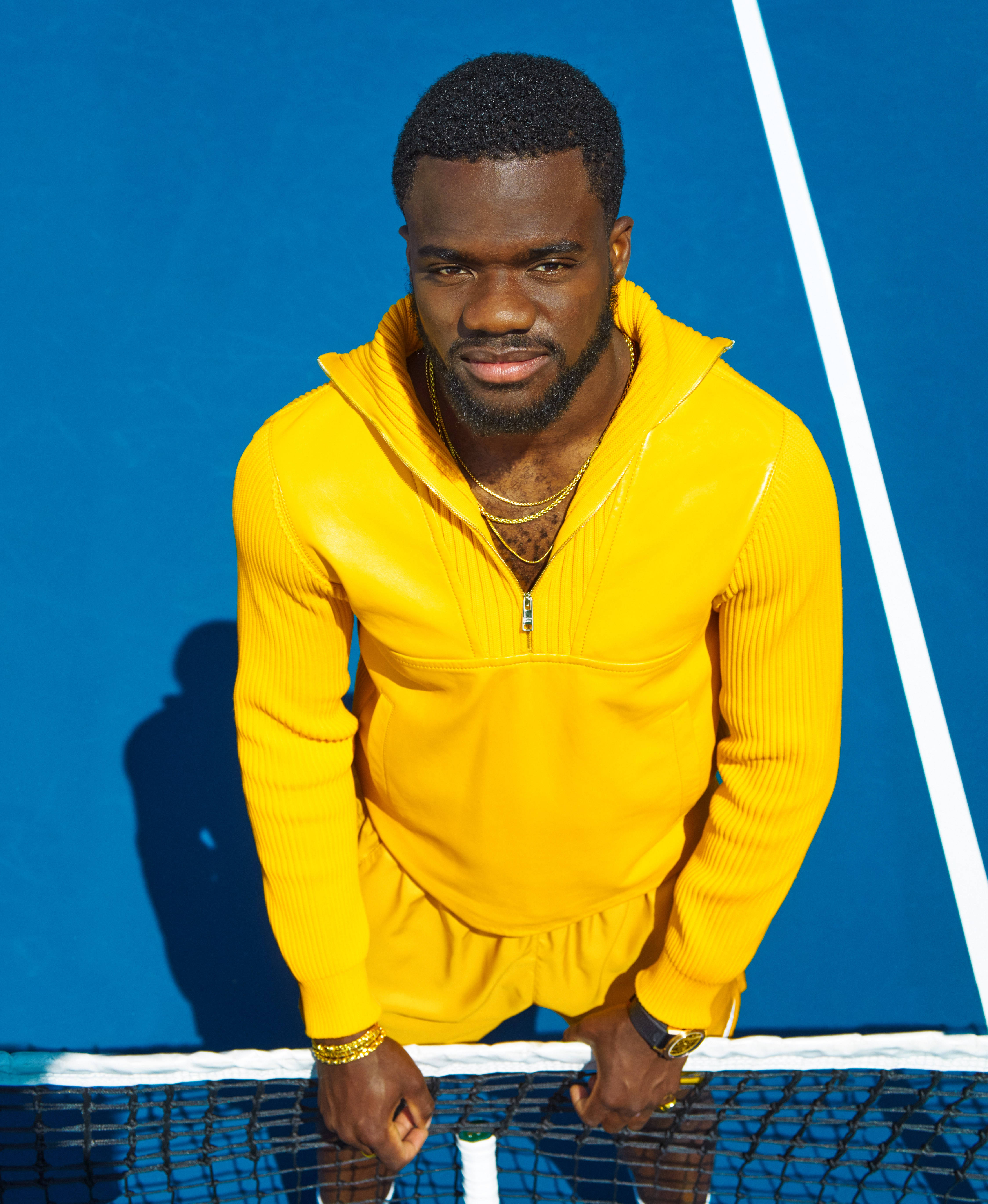 Tennis 桌布 壁紙 — 一名身穿亮黃色羅紋連帽衫的男子在明亮的陽光下，在鮮豔的藍色球場上俯身靠在網球網上。