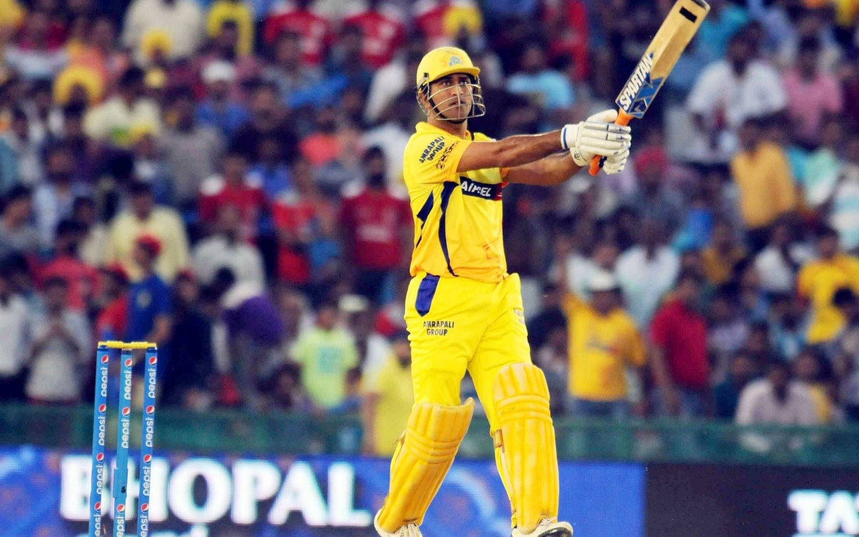 MS Dhoni 桌布 壁紙 — 一名身穿亮黃色裝備的板球擊球手在體育場燈光下的夜間比賽中慶祝強力擊球。
