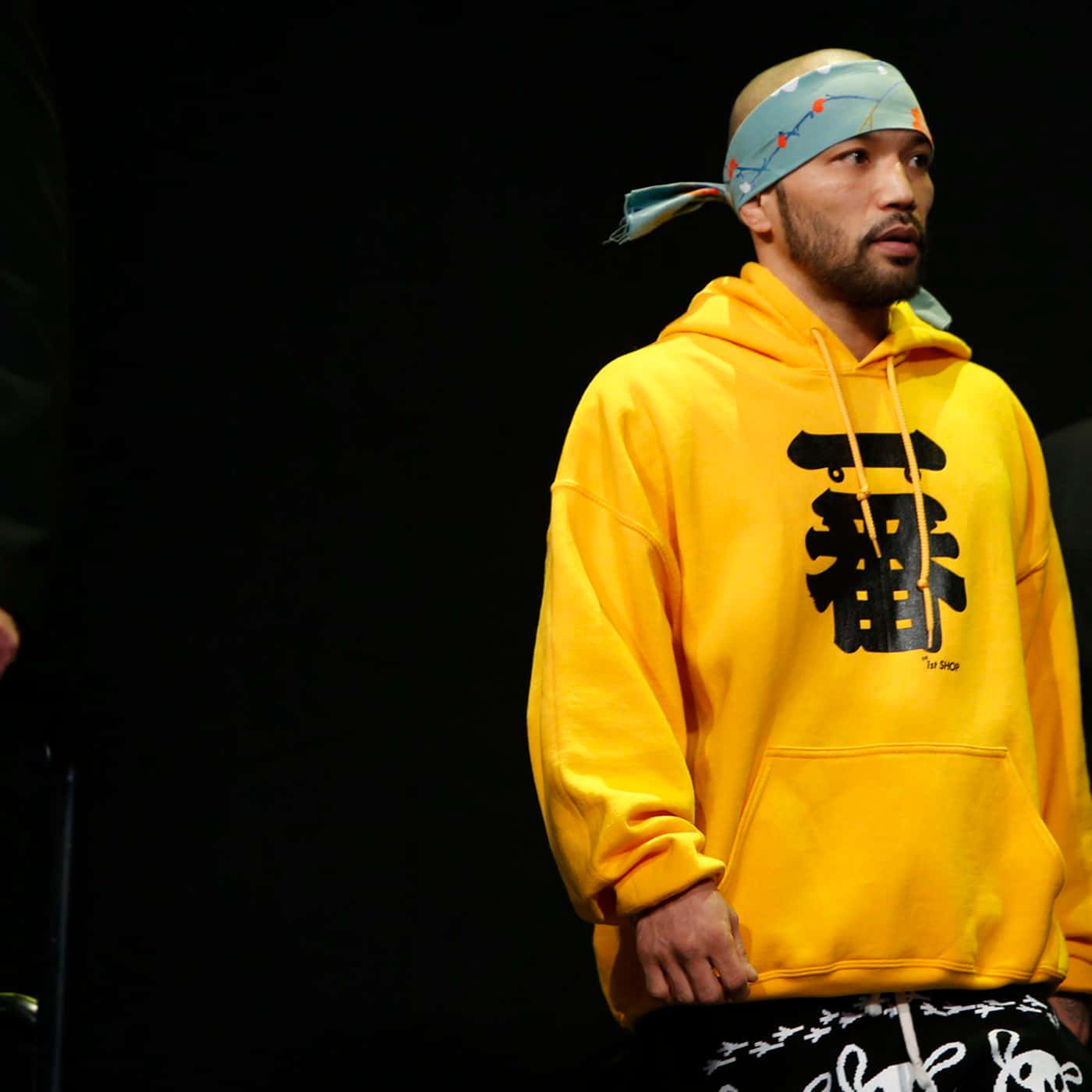 UFC 桌布 壁紙 — 一名身穿亮黃色連帽衫、帶有黑色漢字的男子，戴著圖案頭帶，站在深色背景前。