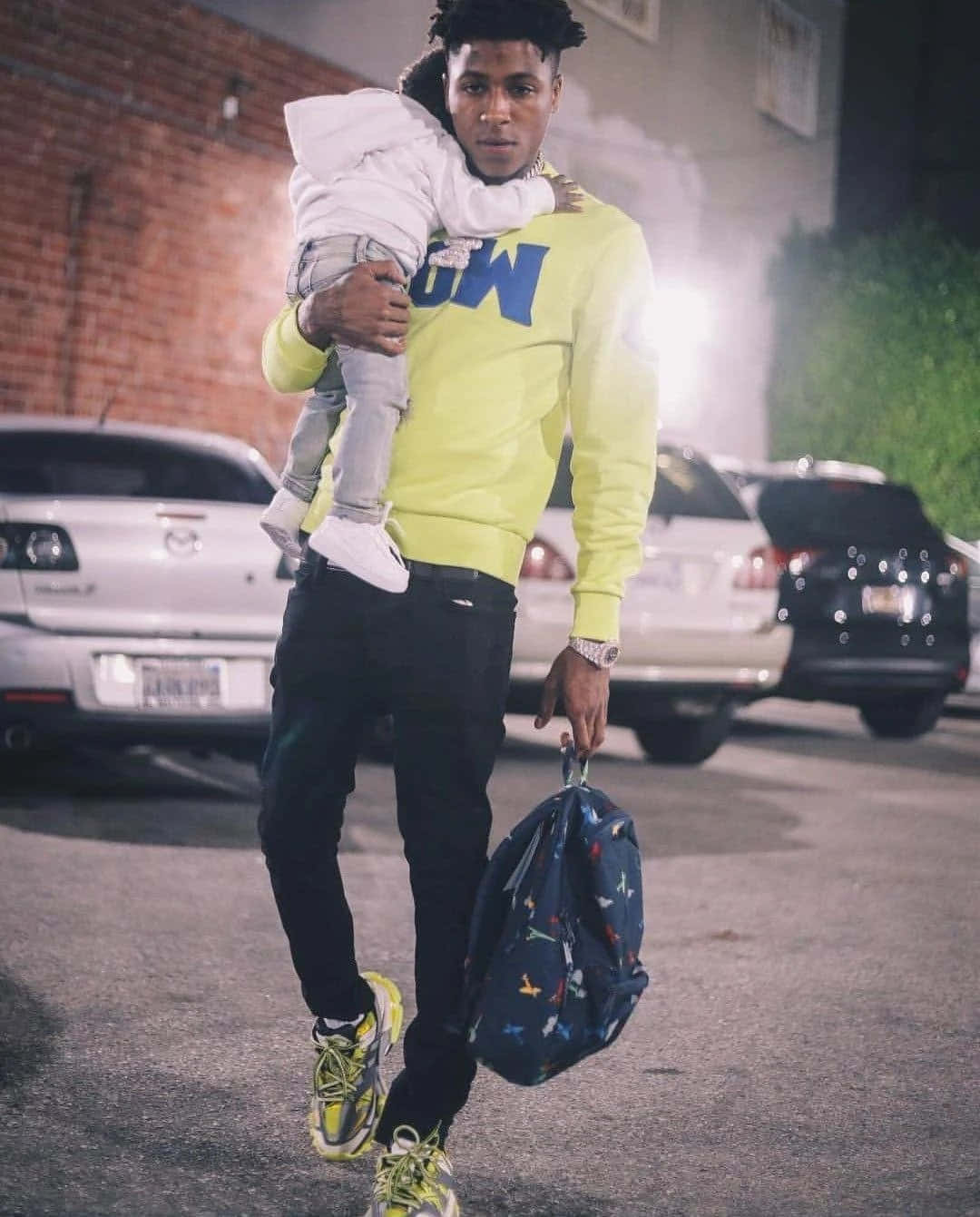 NBA YoungBoy 桌布 壁紙 — 一名身穿亮黃色運動衫的男子在夜間穿過光線昏暗的停車場時抱著一個孩子。