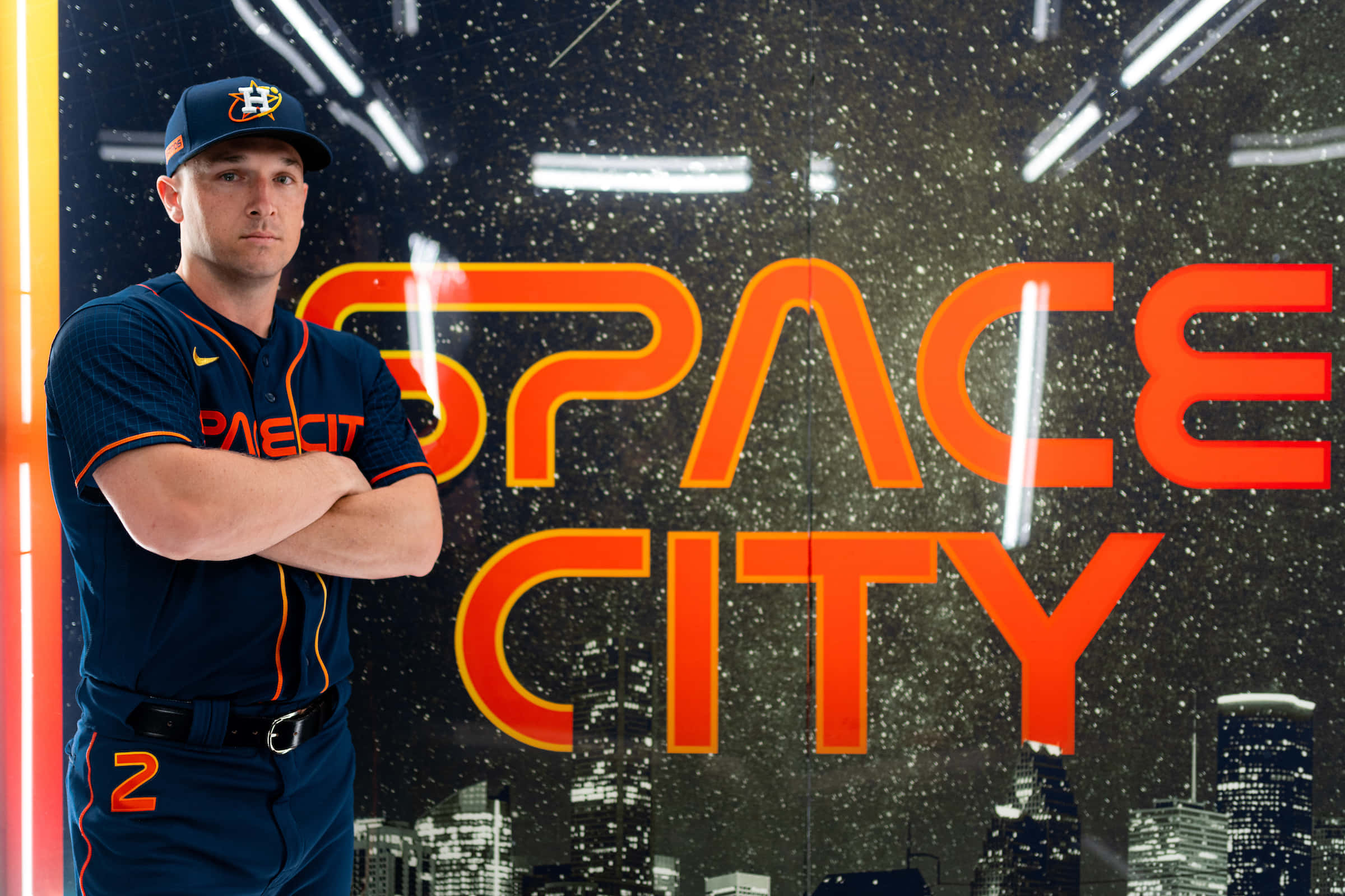Astros 桌布 壁紙 — 一名身穿休士頓太空人隊球衣的棒球運動員，在星光燦爛的城市背景下，自信地交叉雙臂站在發光的「SPACE CITY」標誌前。