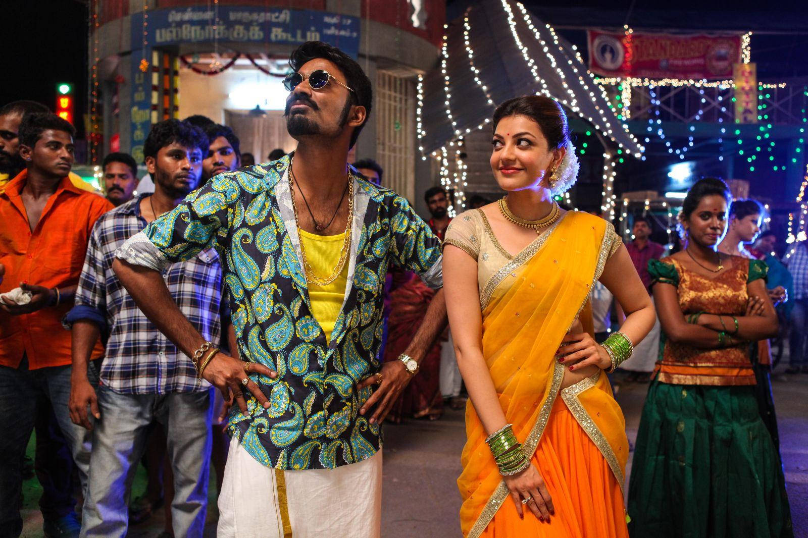 Dhanush 桌布 壁紙 — 一名身穿佩斯利夾克和太陽眼鏡的男子，在夜晚的節日燈光下自信地站在一位身穿黃色紗麗的女子旁邊。