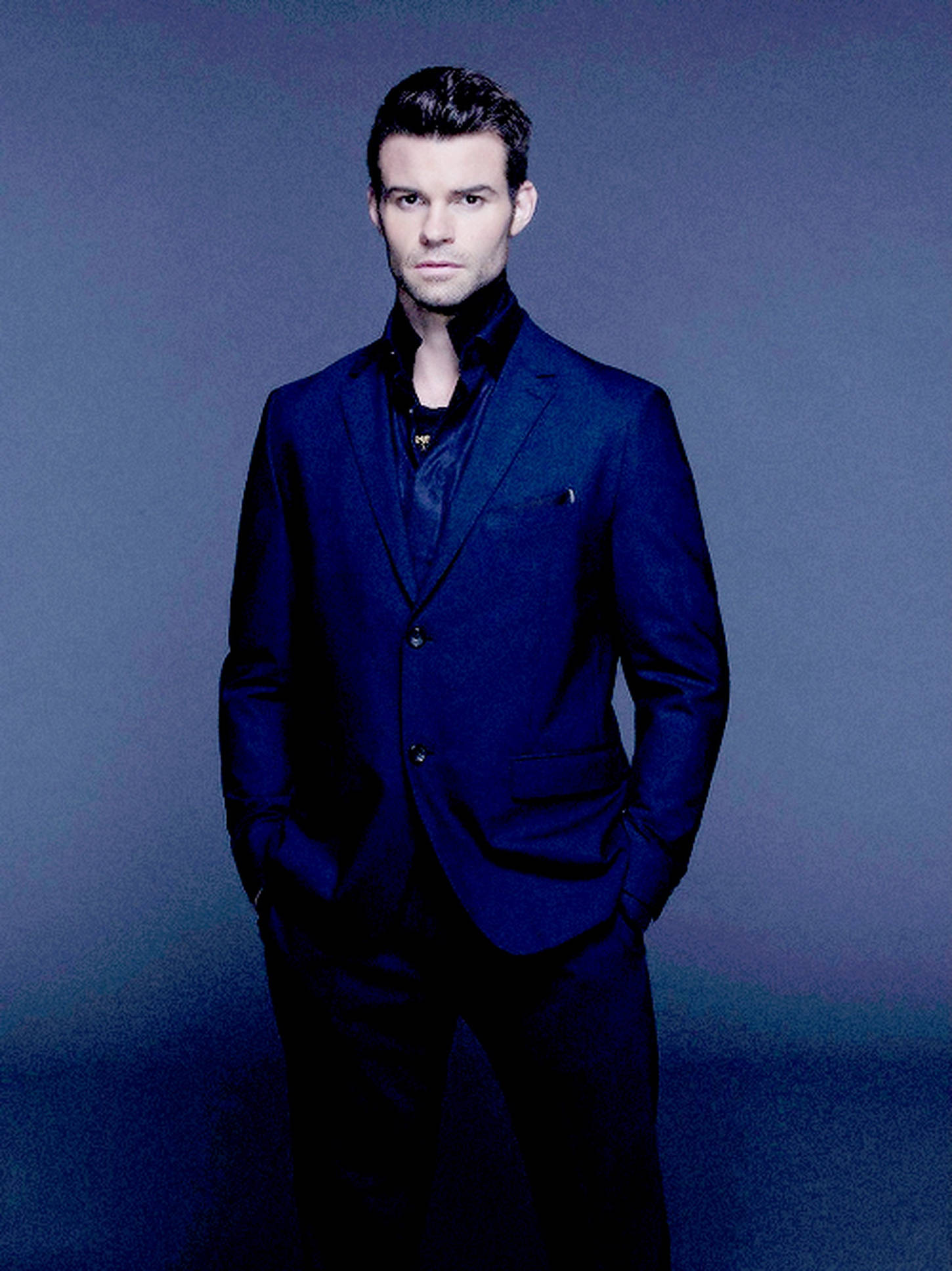 Daniel Gillies 桌布 壁紙 — 一名身穿俐落海軍藍西裝的男子，雙手插袋，自信地站在漸變藍色攝影棚背景前。