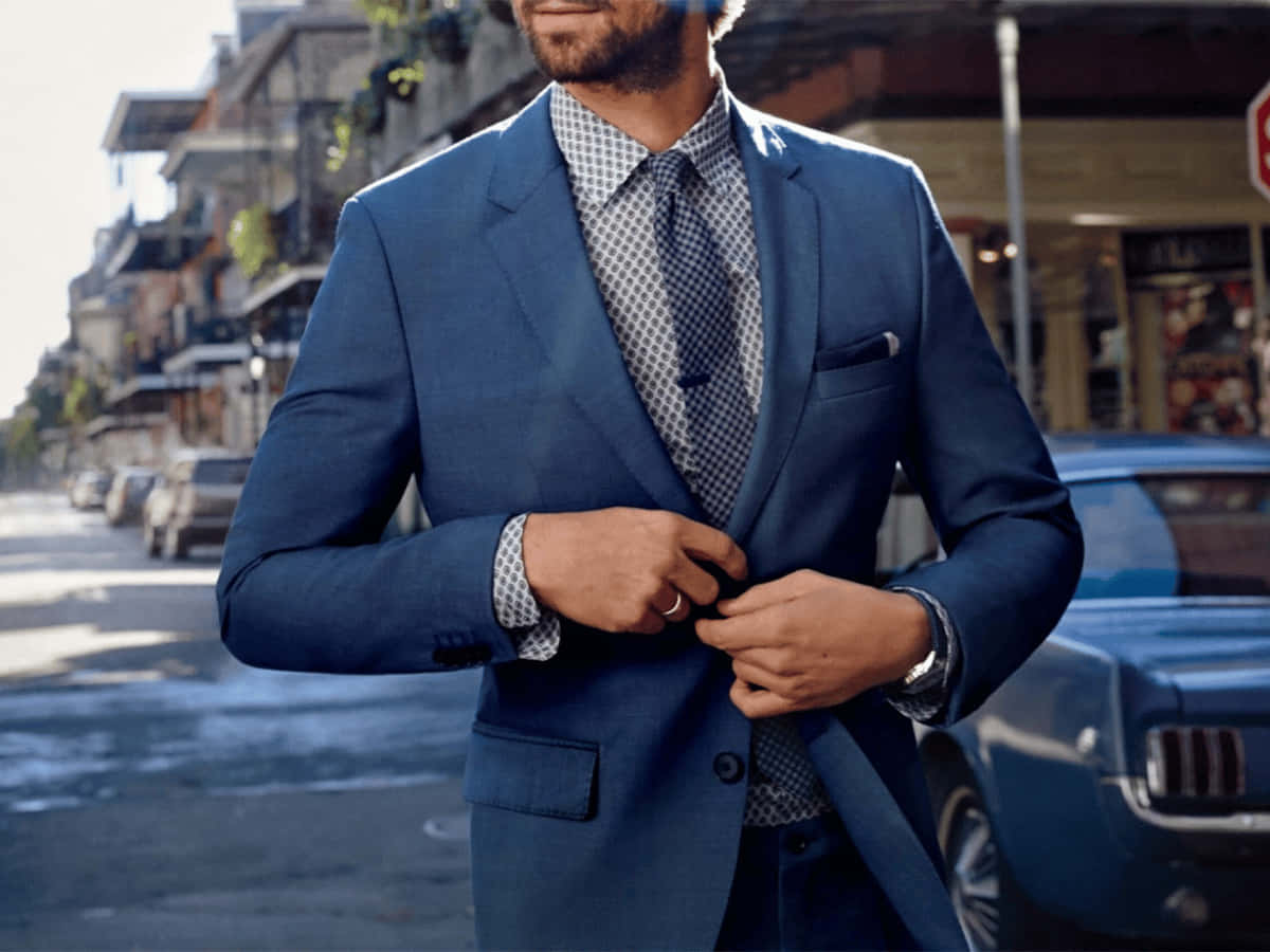 Navy Blue Suit 桌布 壁紙 — 一名身穿俐落藍色西裝的男子，在陽光明媚的城市街道上，在一輛復古藍色汽車旁扣上外套鈕扣。
