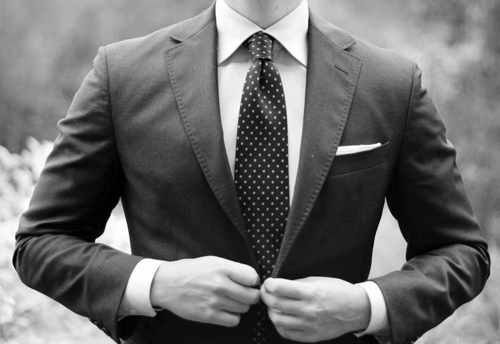 style 桌布 壁紙 — 一名身穿俐落西裝的男子正在扣上外套的鈕扣，雙手清晰可見，背景是單色調模糊的自然景色。