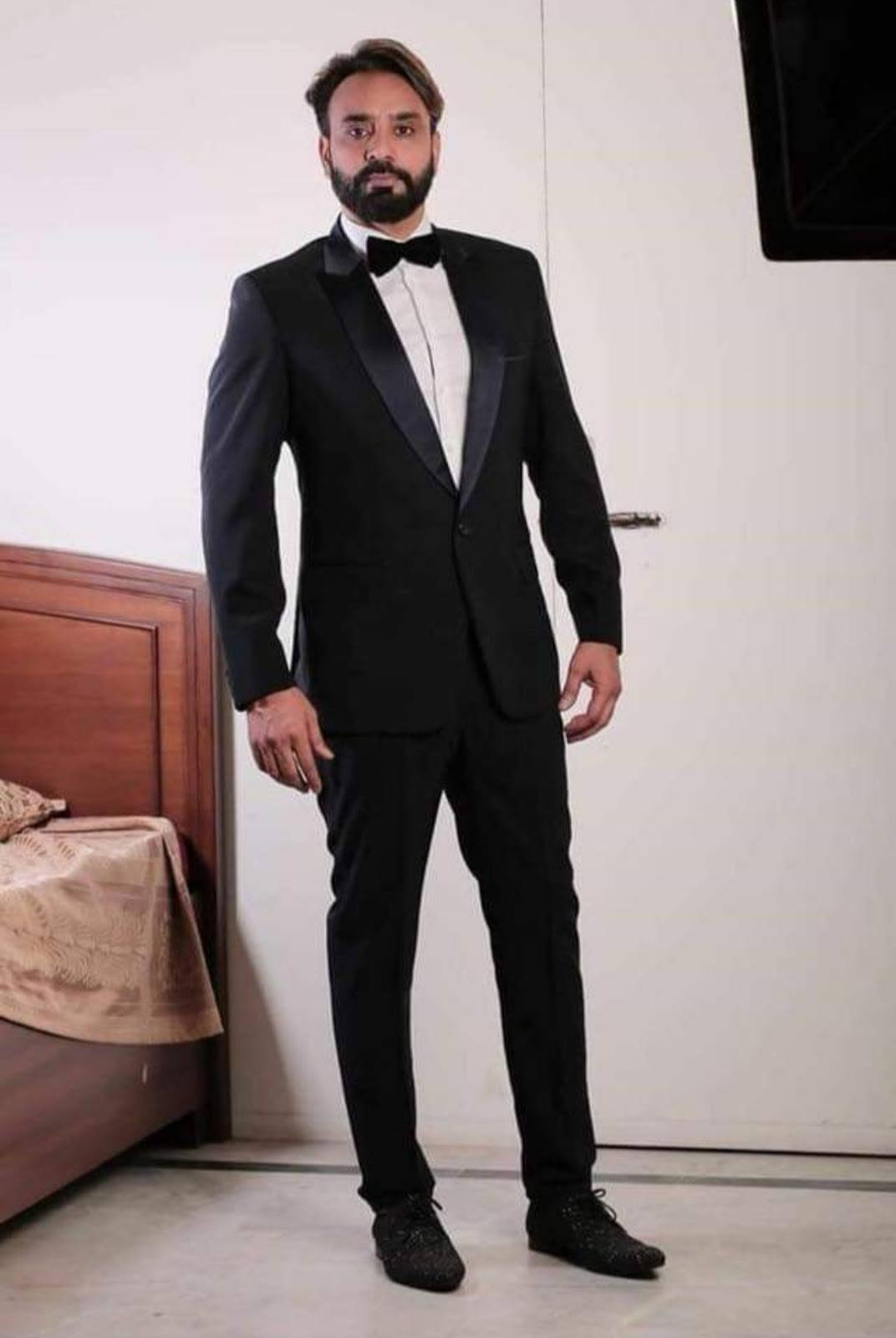 Celebrity 桌布 壁紙 — 一名身穿俐落黑色燕尾服配蝴蝶結的男人，自信地站在一個配有木製床頭板和白色牆壁的極簡主義臥室中。