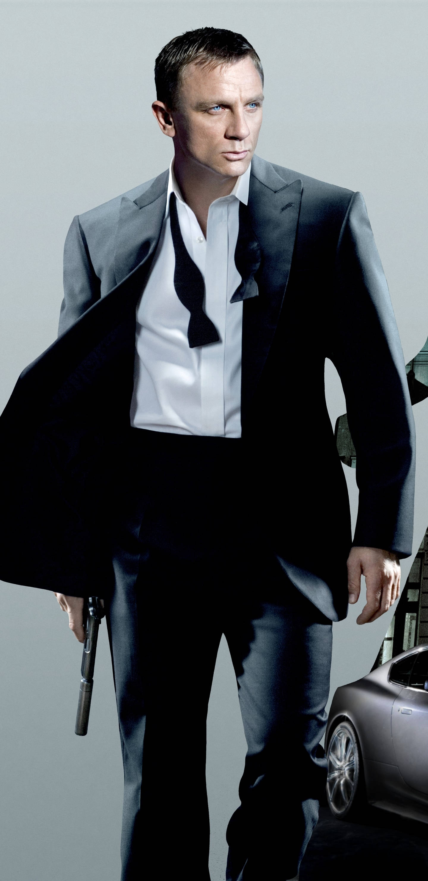 Daniel Craig 桌布 壁紙 — 一名身穿俐落黑色西裝的男子，在純灰色背景前，站在一輛時尚的銀色跑車旁，手持一把手槍。