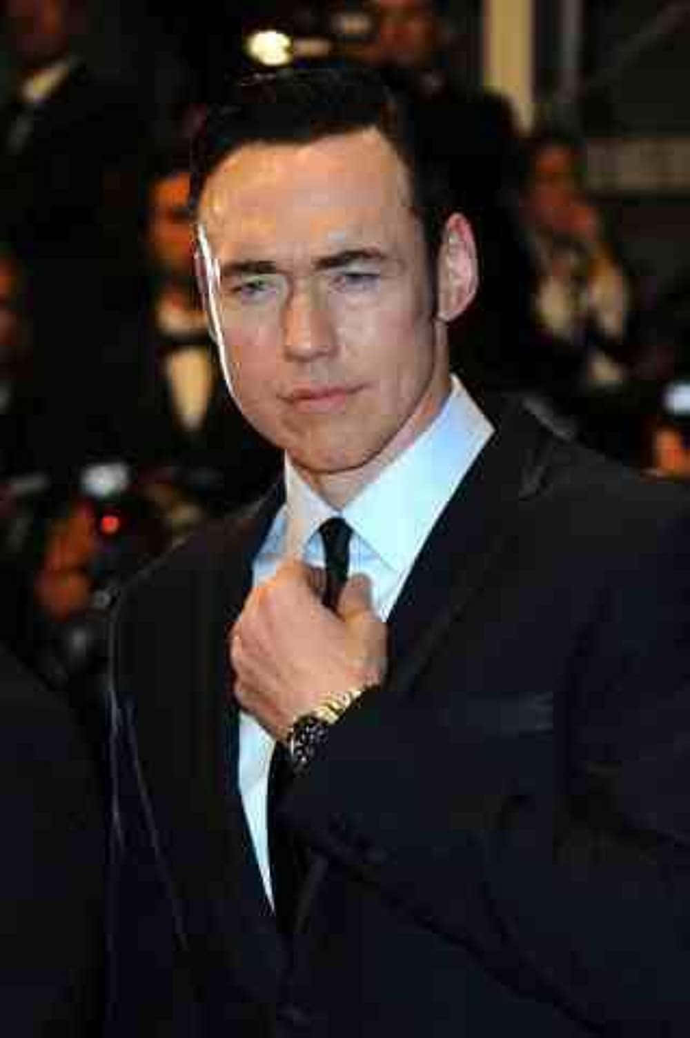 Kevin Durand 桌布 壁紙 — 一名身穿俐落黑色西裝的男子，在閃光燈下於華麗的紅地毯活動中調整領帶。