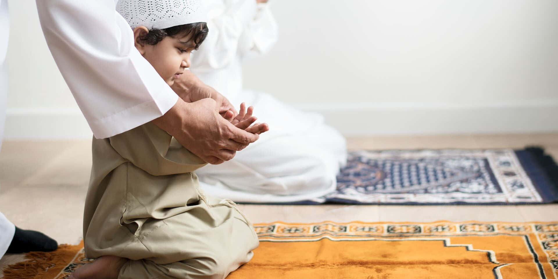 Praying 桌布 壁紙 — 一名身穿傳統伊斯蘭服飾的男孩在室內有圖案的祈禱毯上，跪在成年人旁邊祈禱。