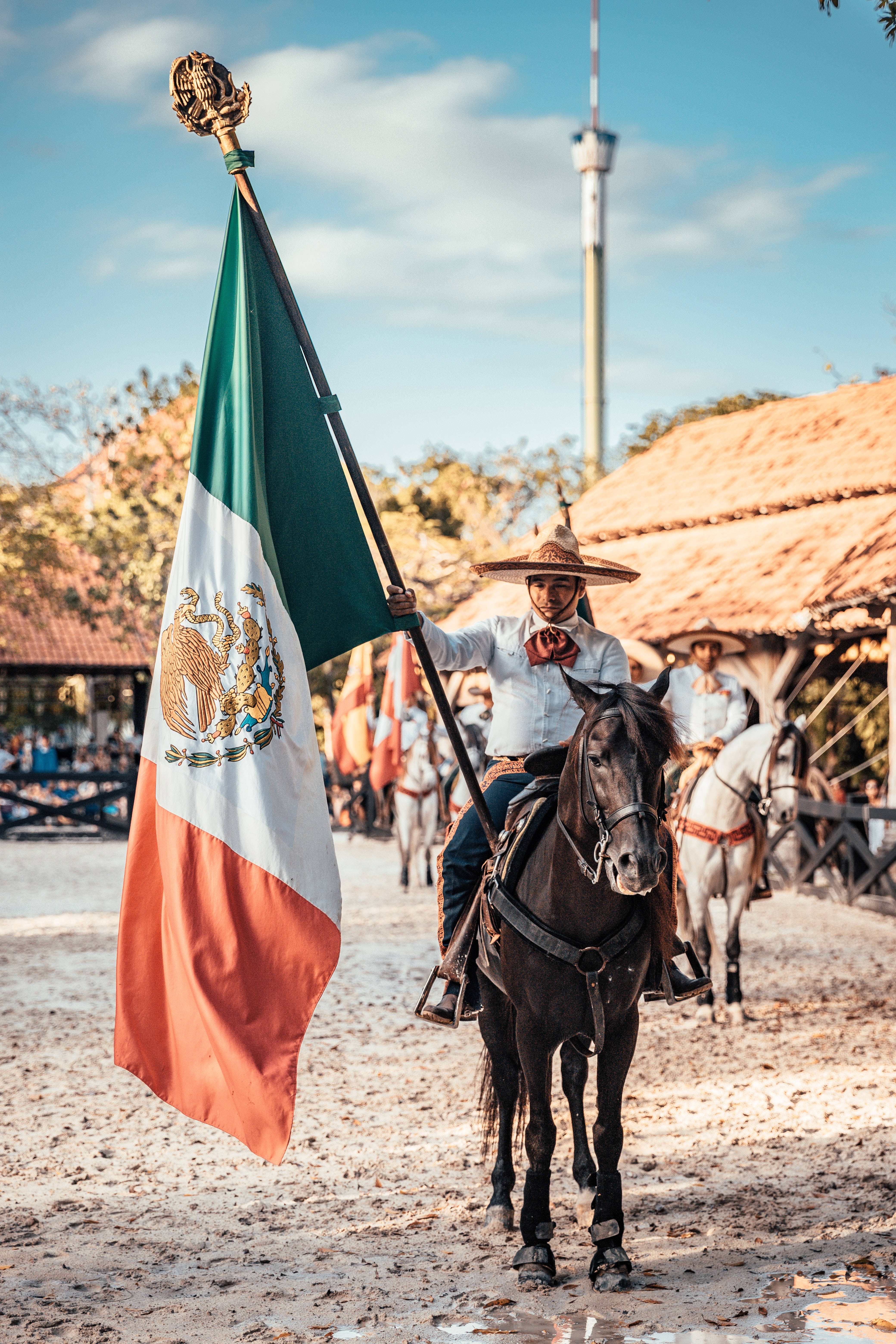 Mexican 桌布 壁紙 — 一名身穿傳統墨西哥查羅服飾的騎手，騎著一匹黑馬，在陽光明媚、塵土飛揚的閱兵場上自豪地高舉國旗。