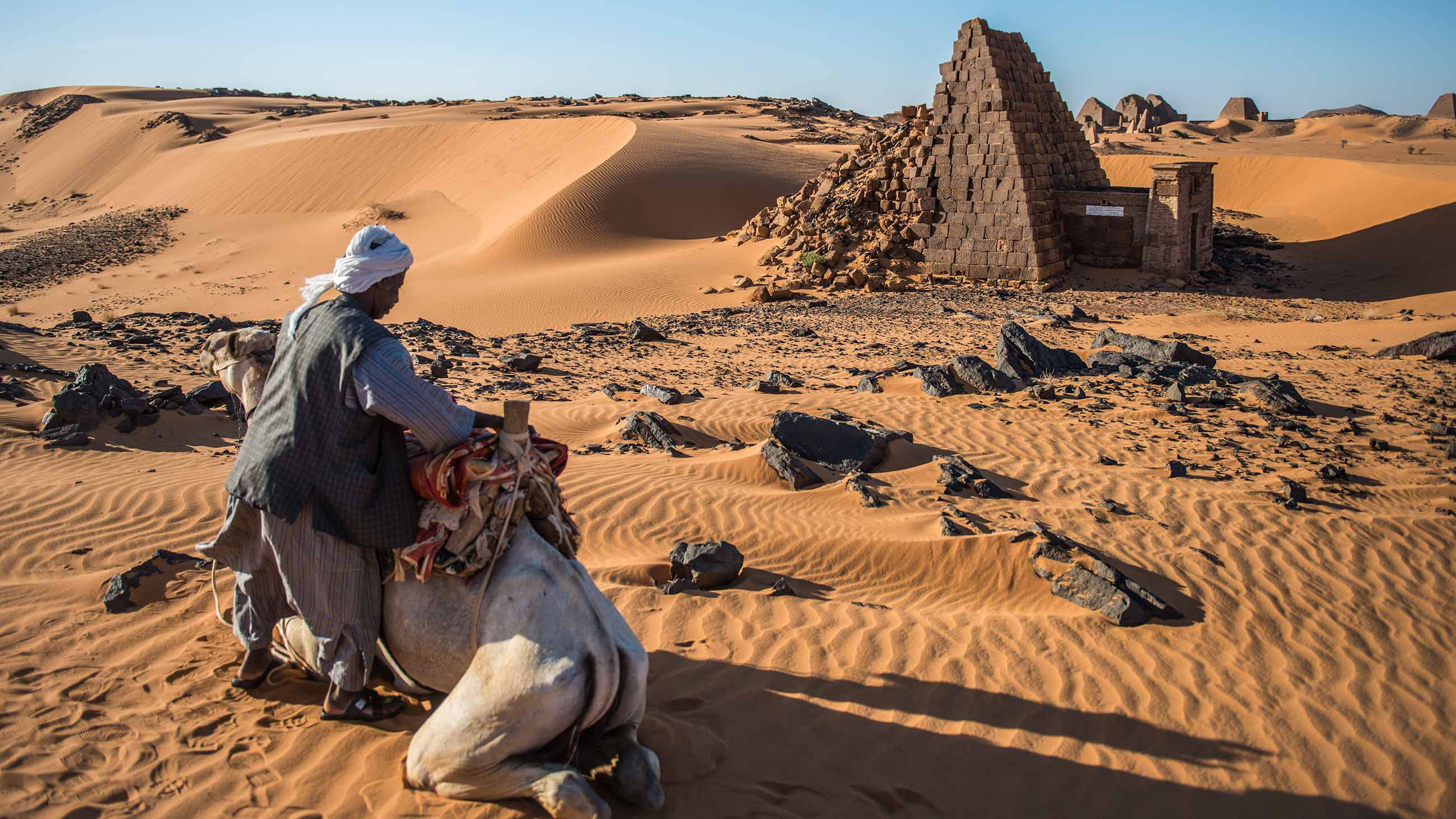 Sudan 桌布 壁紙 — 一名身穿傳統服飾的男子騎著一頭跪著的駱駝，在晴朗的藍天下穿越金色沙丘，靠近一座風化的石頭廢墟。