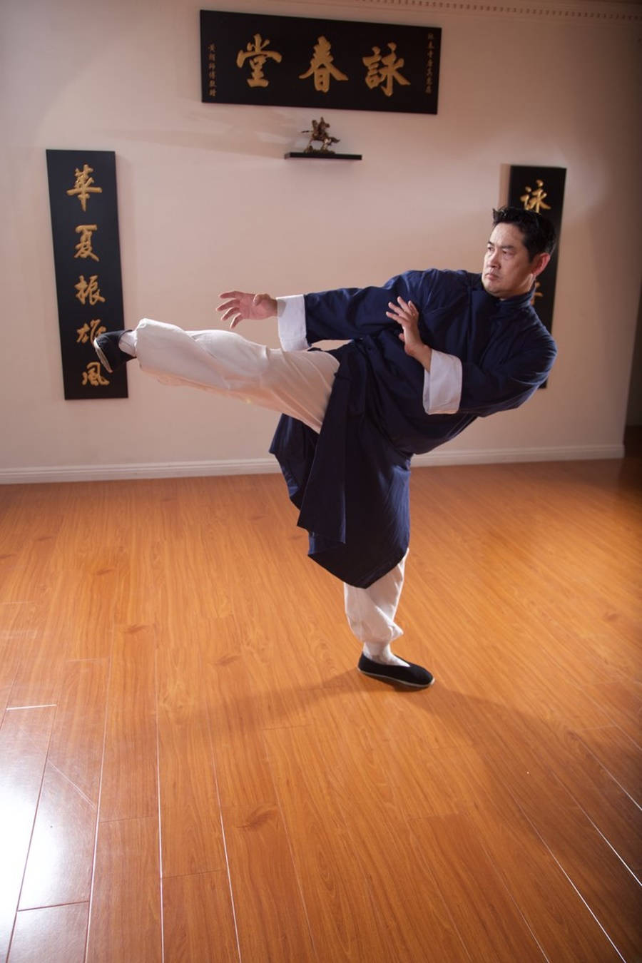 Kung Fu 桌布 壁紙 — 一名身穿傳統武術服裝的男子，在裝飾有中國書法橫幅的木地板工作室中表演高踢腿。