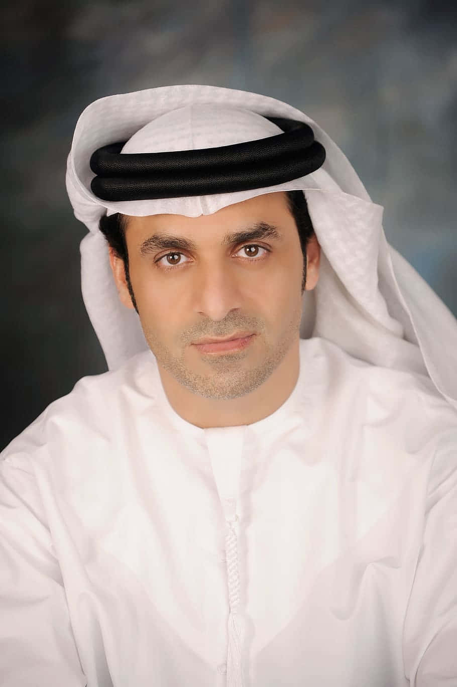 Arabia 桌布 壁紙 — 一名身穿傳統白色坎杜拉和古特拉、戴著黑色阿加爾的男子，在柔和模糊的背景前直視鏡頭。