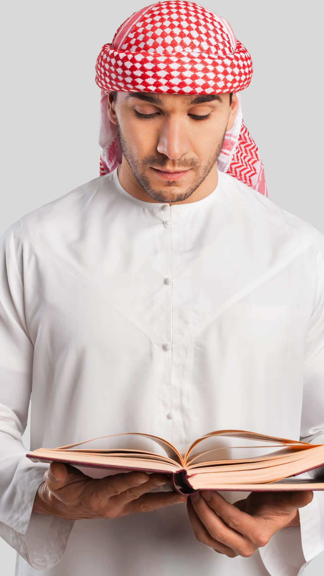 muslim 桌布 壁紙 — 一名身穿傳統紅白格紋頭飾和白色長袍的男子，在純灰色背景下專注而虔誠地閱讀一本書。