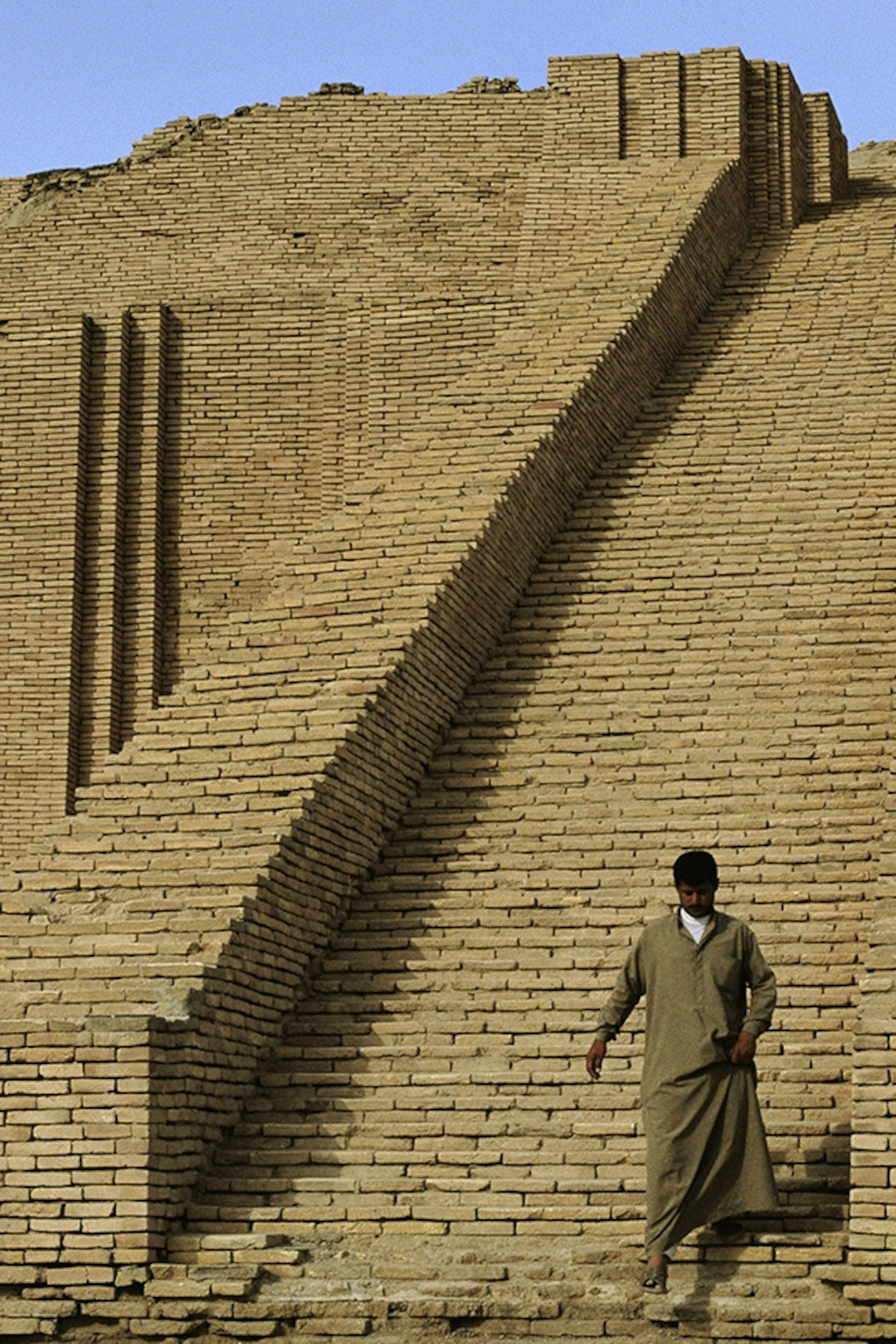 Iraq 桌布 壁紙 — 一名身穿傳統長袍的男子在晴朗的藍天下，沿著古老金字形神塔陡峭的陽光照射的磚坡道走下來。