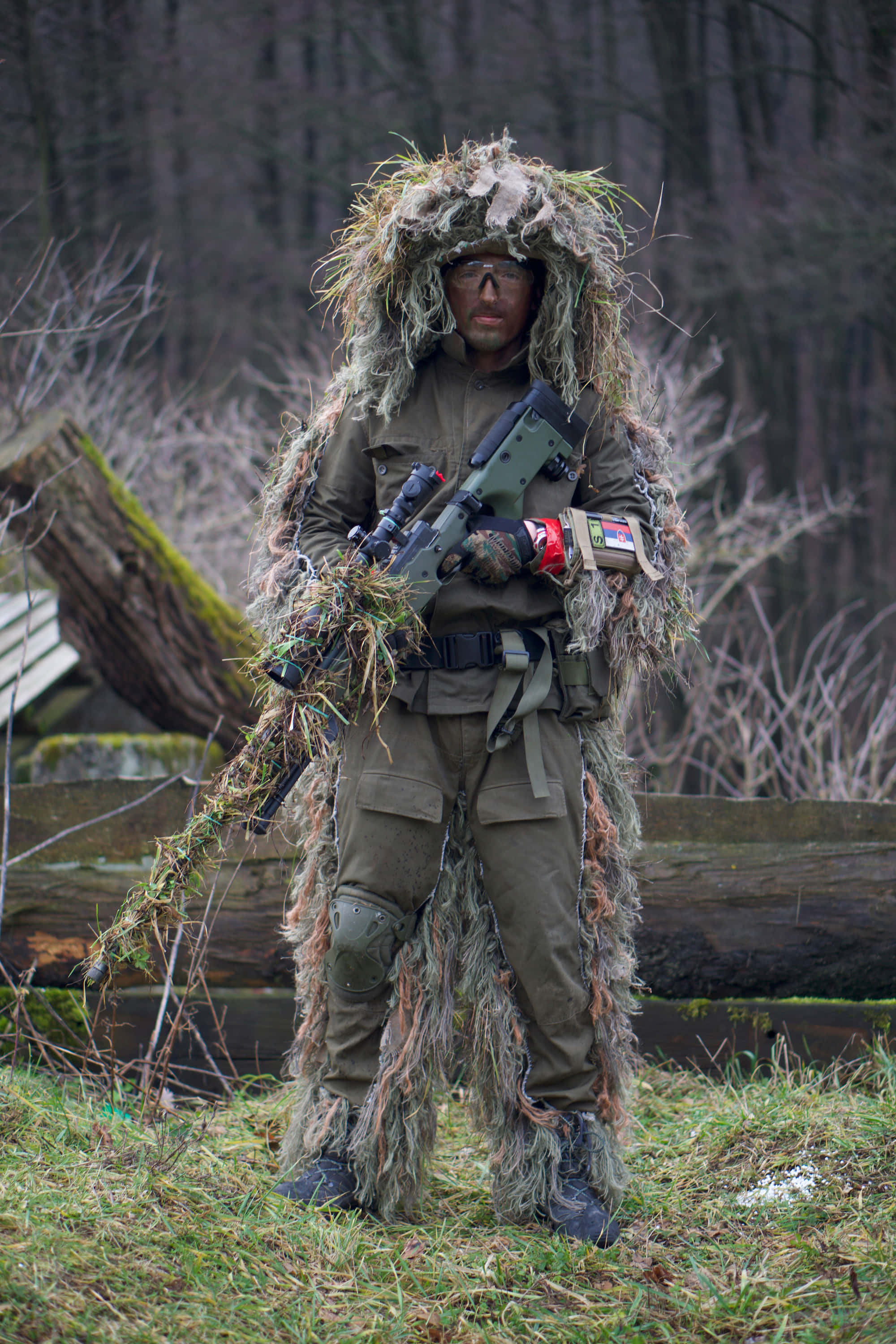 soldier 桌布 壁紙 — 一名身穿全套吉利服和護目鏡的士兵在長滿苔蘚的森林中警覺地站立，手持帶瞄準鏡的步槍。