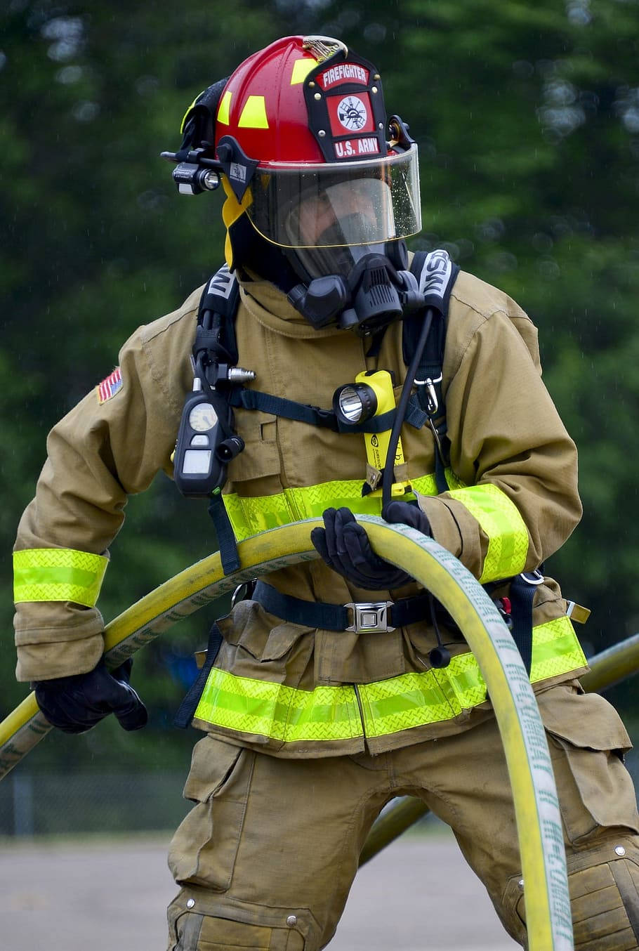 Fireman 桌布 壁紙 — 一名身穿全套裝備的消防員，在模糊的綠色森林背景下抓著一條黃色水帶。