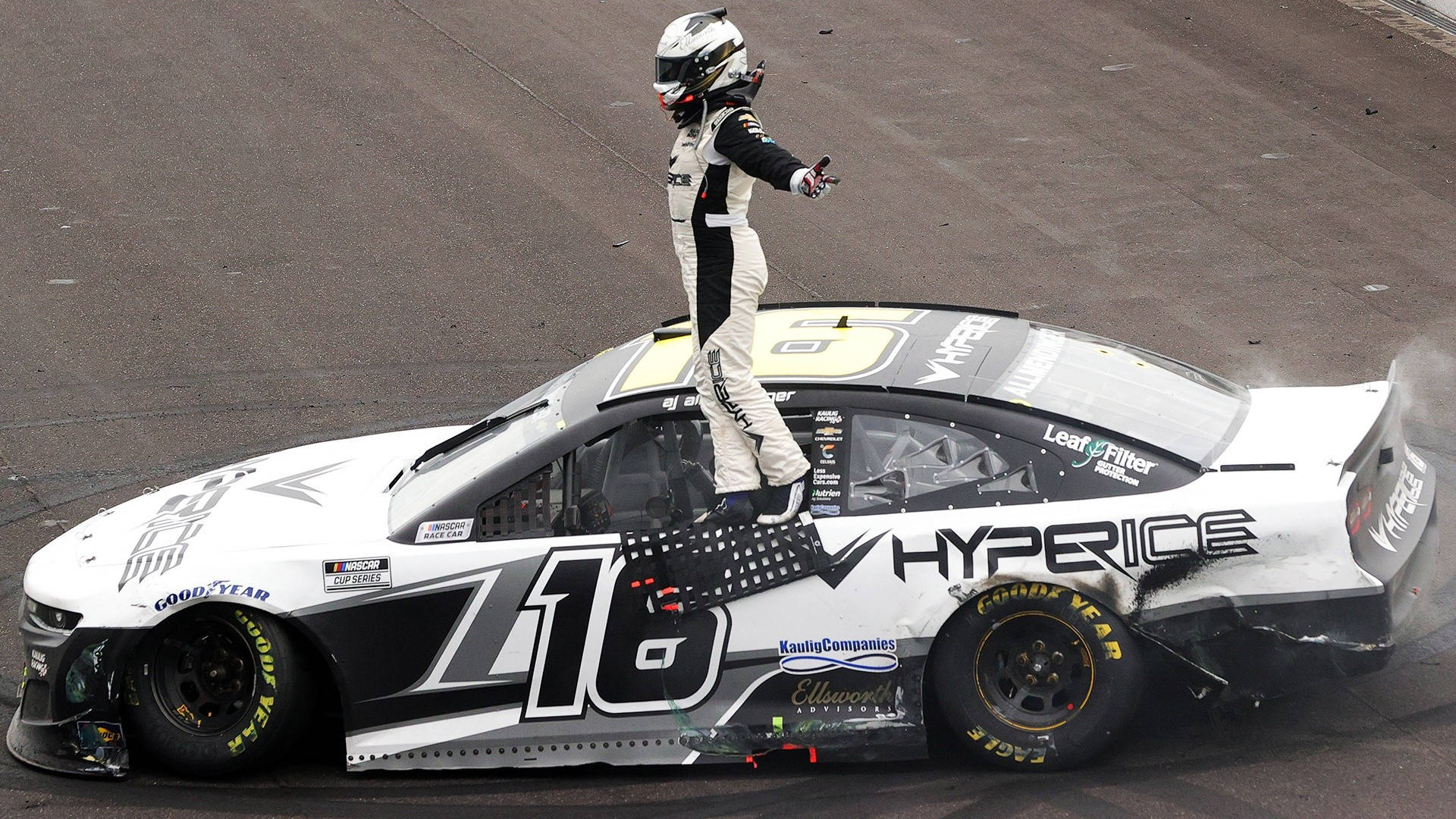 跑車 桌布 壁紙 — 一名身穿全套裝備的賽車手勝利地站在他受損的#16 NASCAR賽車車頂上，賽道上後輪胎冒出滾滾濃煙。