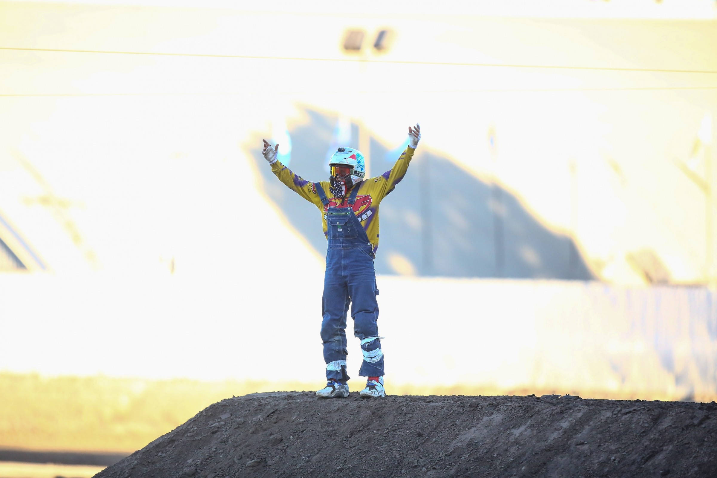 Ronnie Mac 桌布 壁紙 — 一名身穿全套裝備的越野摩托車手，在明亮的陽光背景下，站在土堆頂上高舉雙臂，展現勝利姿態。