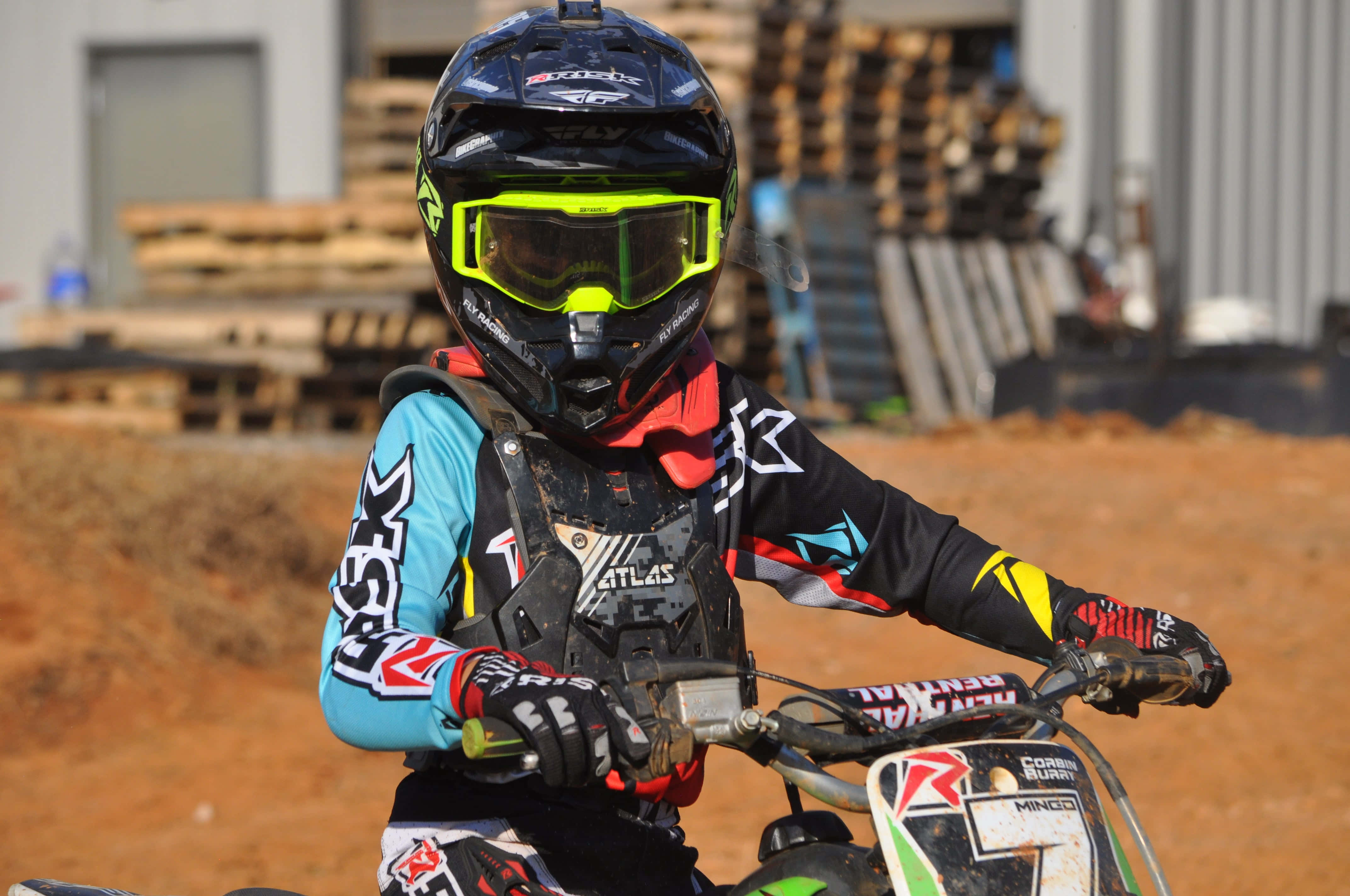 Dirtbike 桌布 壁紙 — 一名身穿全套裝備的越野摩托車手在塵土飛揚的賽道上緊握 dirt bike 的把手，背景有堆疊的木製托盤。