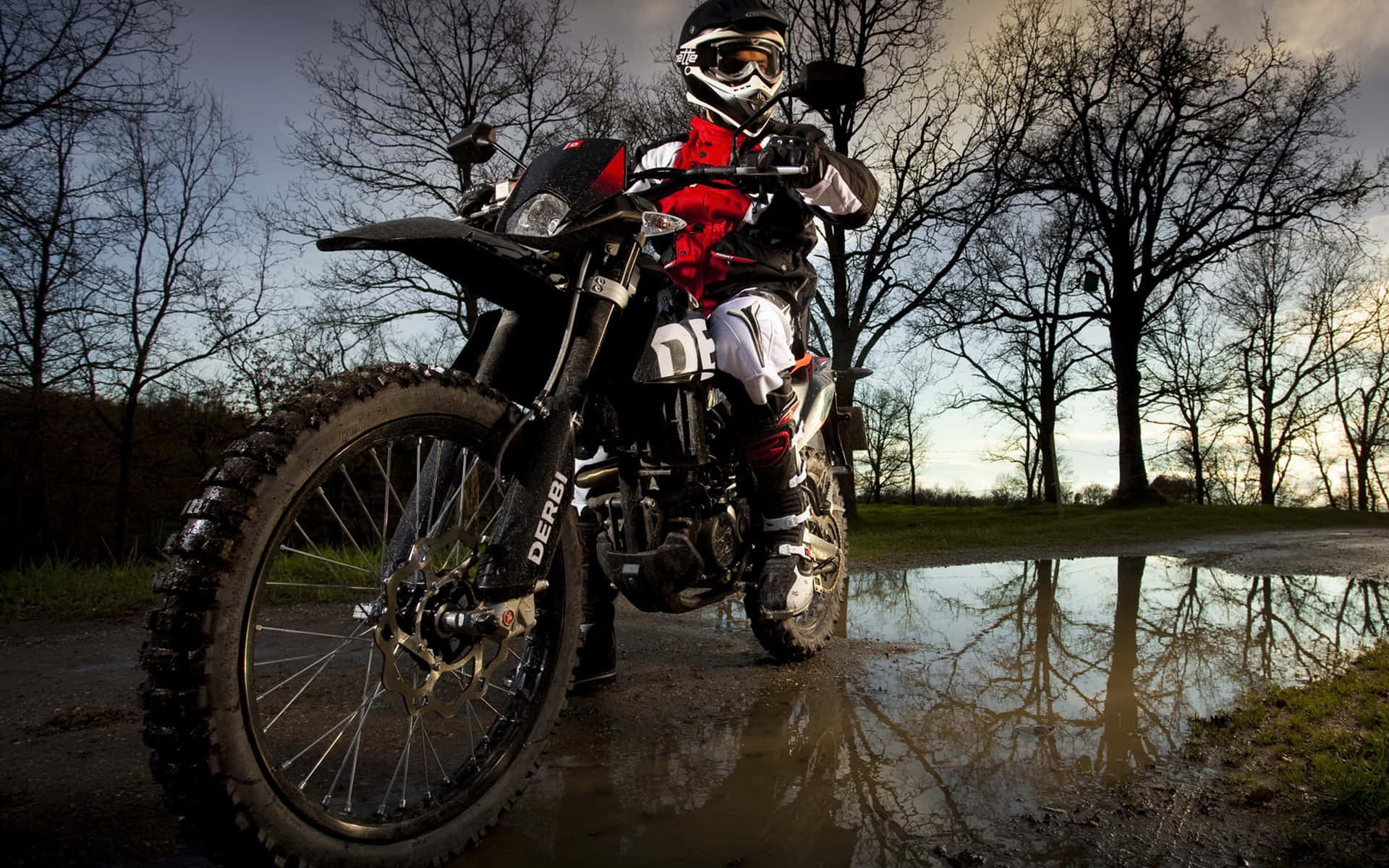 Motocross 桌布 壁紙 — 一名身穿全套裝備的越野摩托車騎士，在陰沉天空下，站在堅固的越野摩托車旁，旁邊是一個反射的水坑。