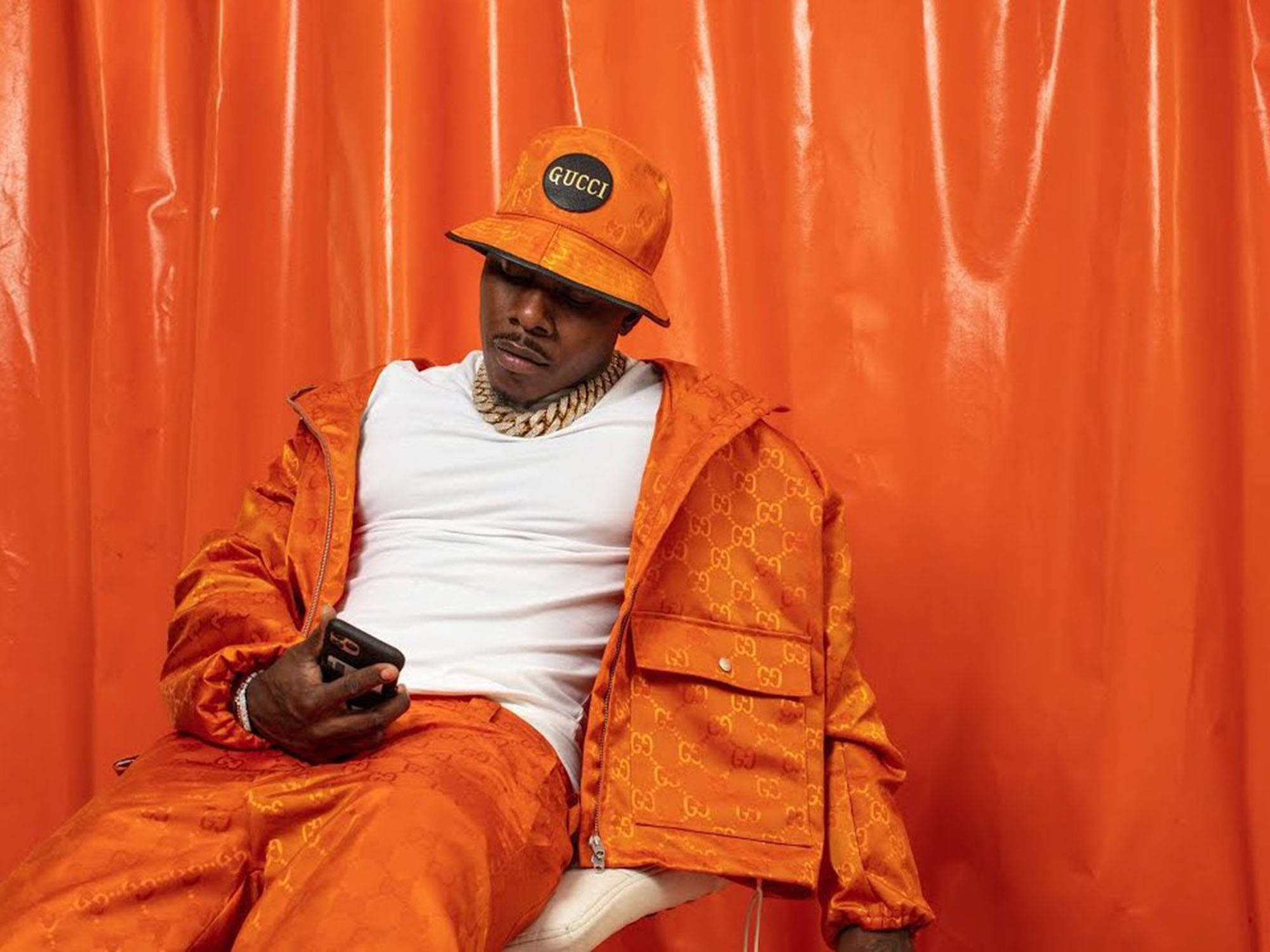 Dababy 桌布 壁紙 — 一名身穿全橙色Gucci服裝的男子，靠著相配的橙色窗簾，放鬆地坐著，手持電話。