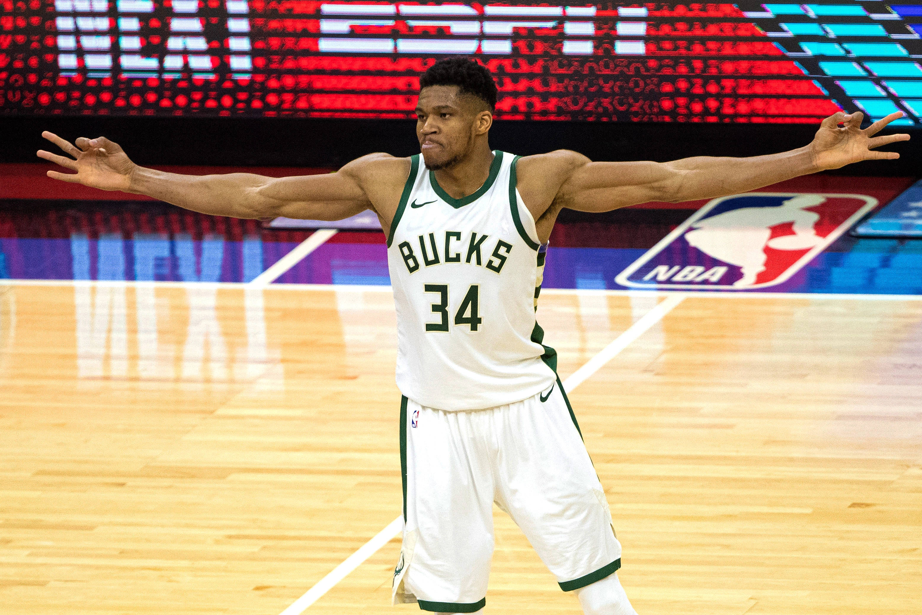 Giannis 桌布 壁紙 — 一名身穿公鹿隊球衣的籃球運動員在明亮的場館燈光下，在拋光球場上張開雙臂慶祝。