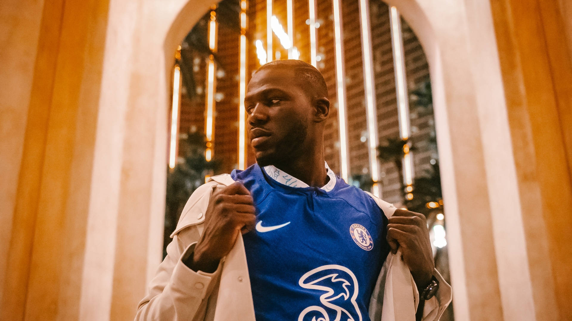 Athletic 桌布 壁紙 — 一名身穿切爾西FC球衣的男子在溫暖的金色拱門下拉開他的米色外套，背後是城市燈光。