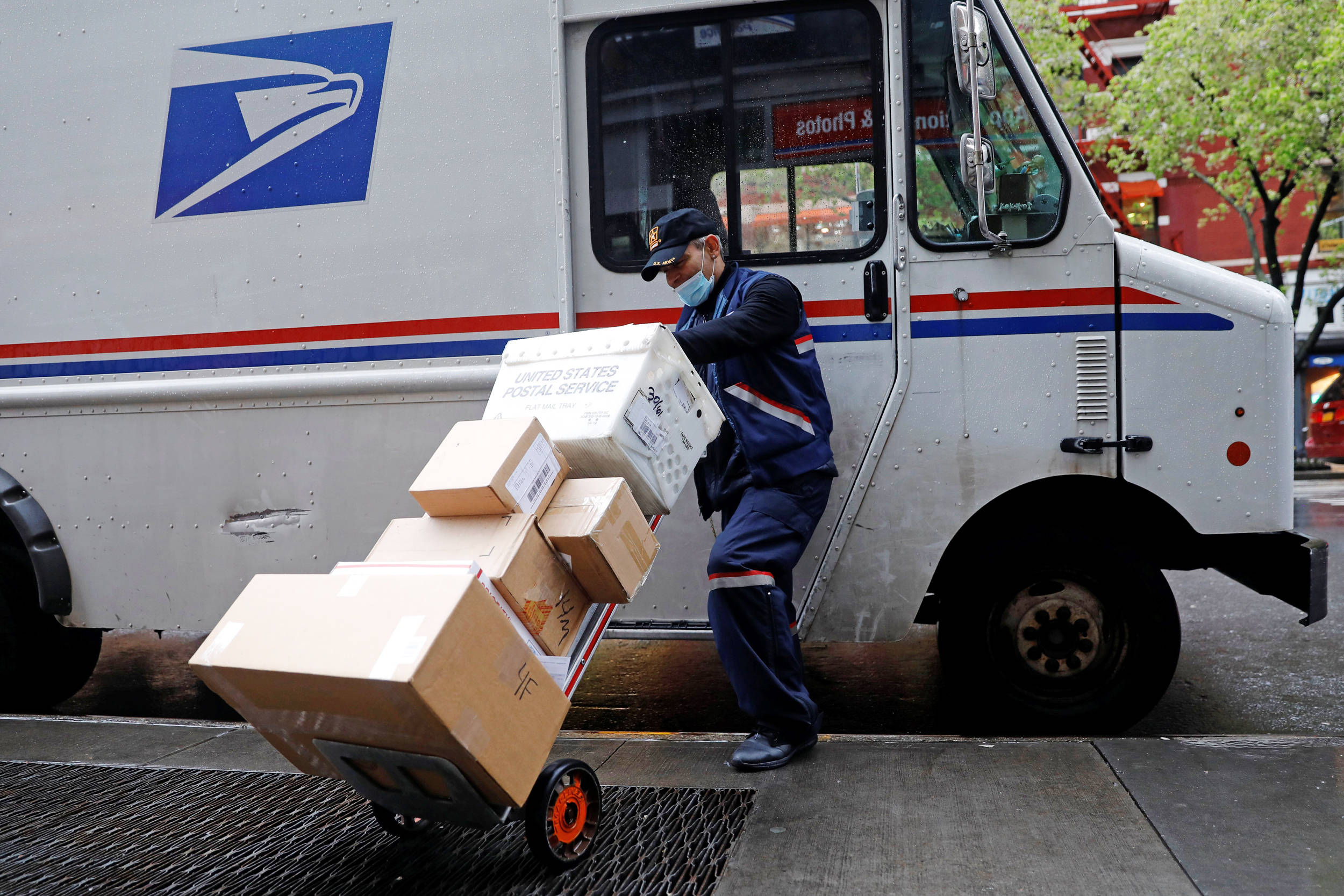 Box 桌布 壁紙 — 一名身穿制服及口罩的美國郵政（USPS）郵差，在濕滑的城市人行道上，將沉重包裹裝載到白色郵車旁的推車上。