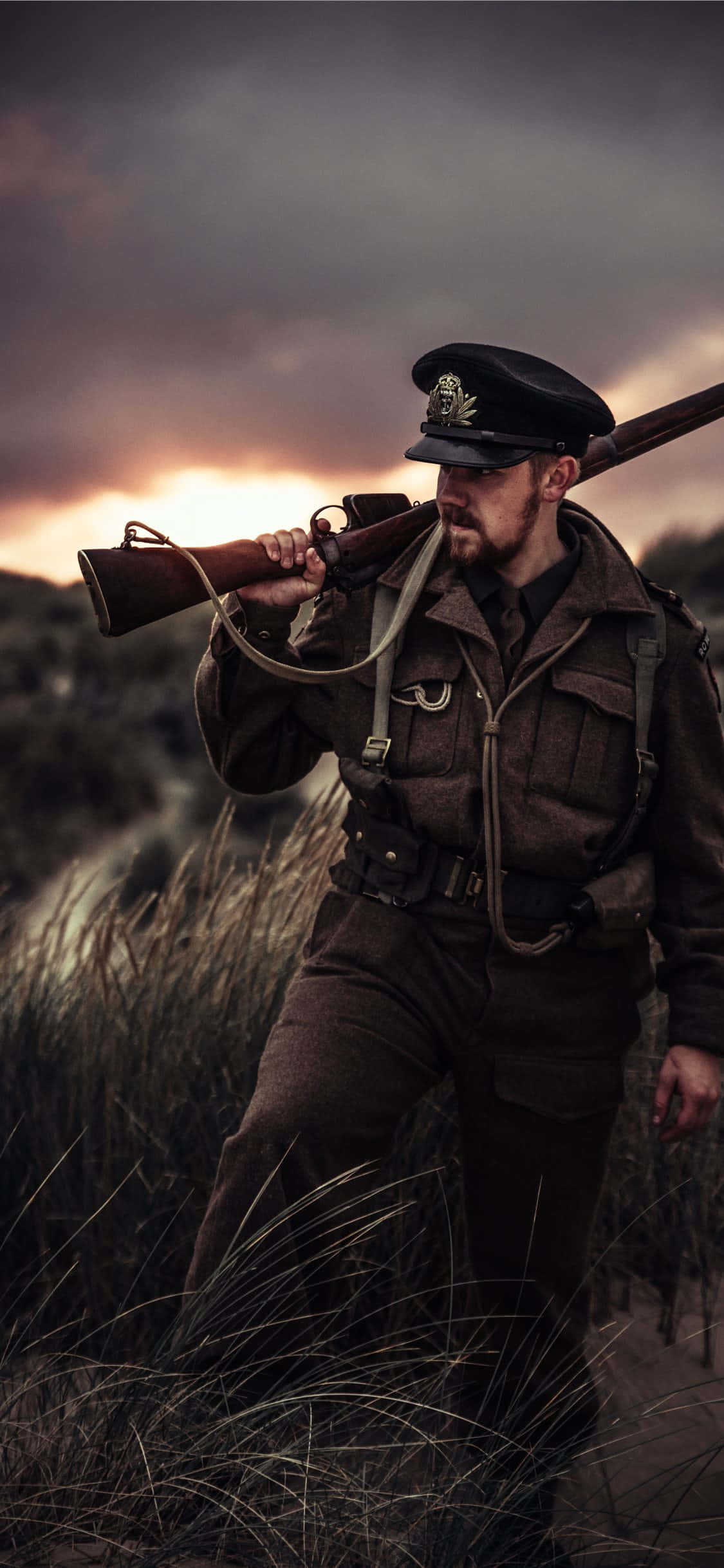 Ww2 iPhone 桌布 壁紙 — 一名身穿制服的士兵在戲劇性的暮色天空下，背著步槍穿過高草叢。