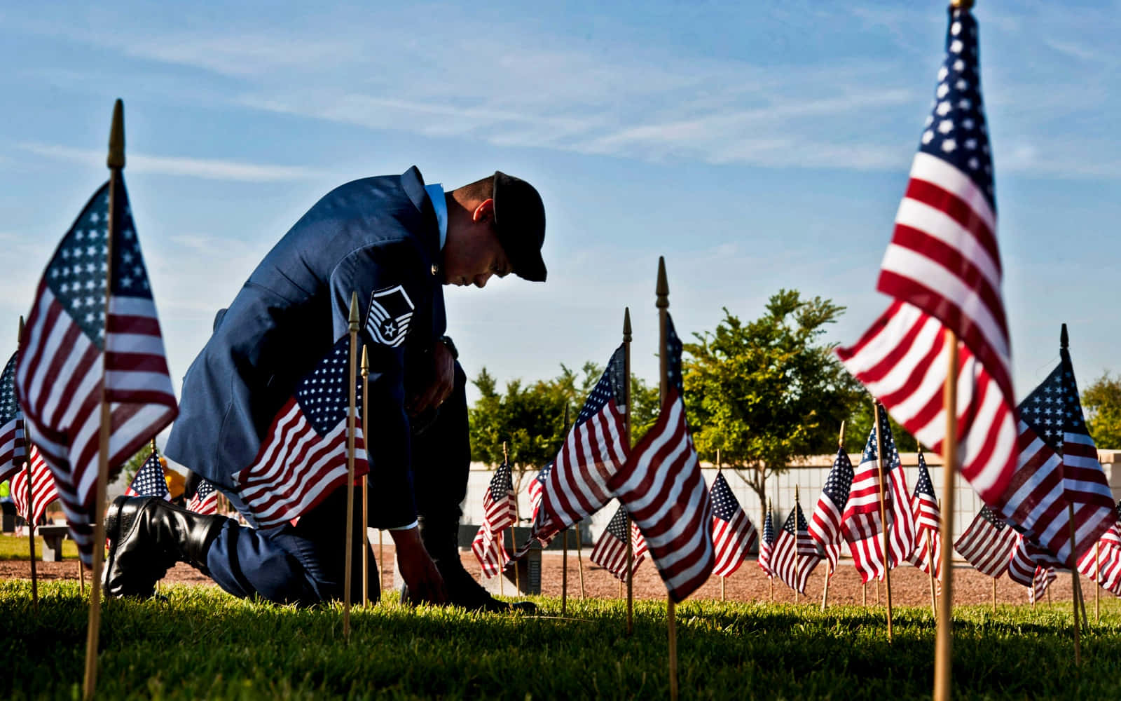 Memorial Day 桌布 壁紙 — 一名身穿制服的軍人莊嚴地跪在草地上，在晴朗的藍天下於紀念地點放置美國國旗。