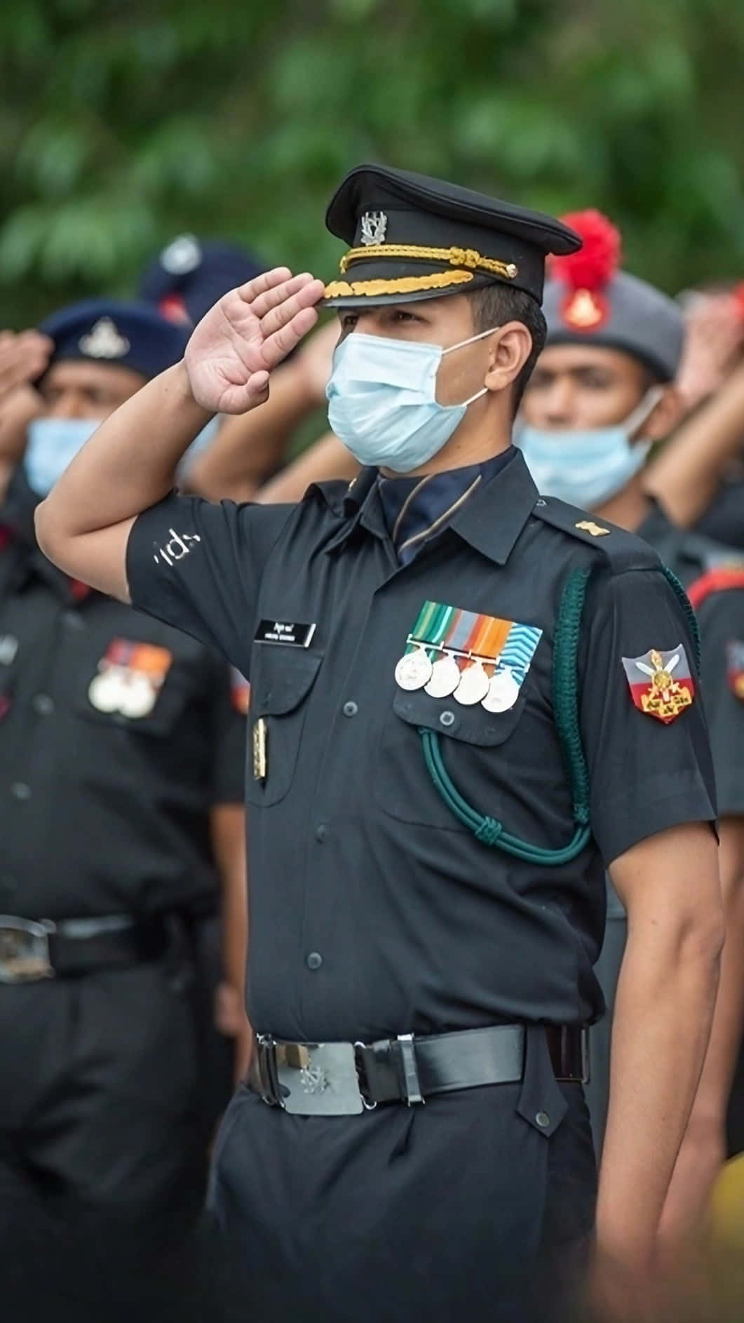 印度軍隊 桌布 壁紙 — 一名身穿制服的軍官在戶外正式儀式上戴著口罩敬禮，背景有模糊的同事。