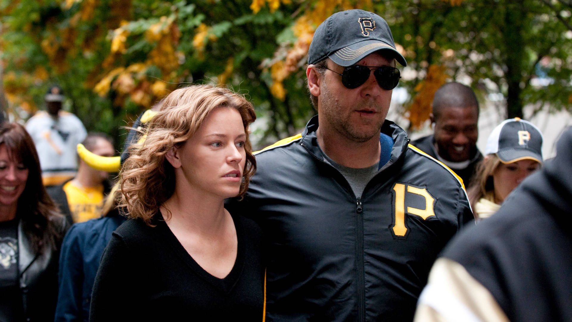 Russell Crowe 桌布 壁紙 — 一名身穿匹茲堡海盜隊外套和帽子的男子，在秋天的樹下與一名女子一起穿過人群。