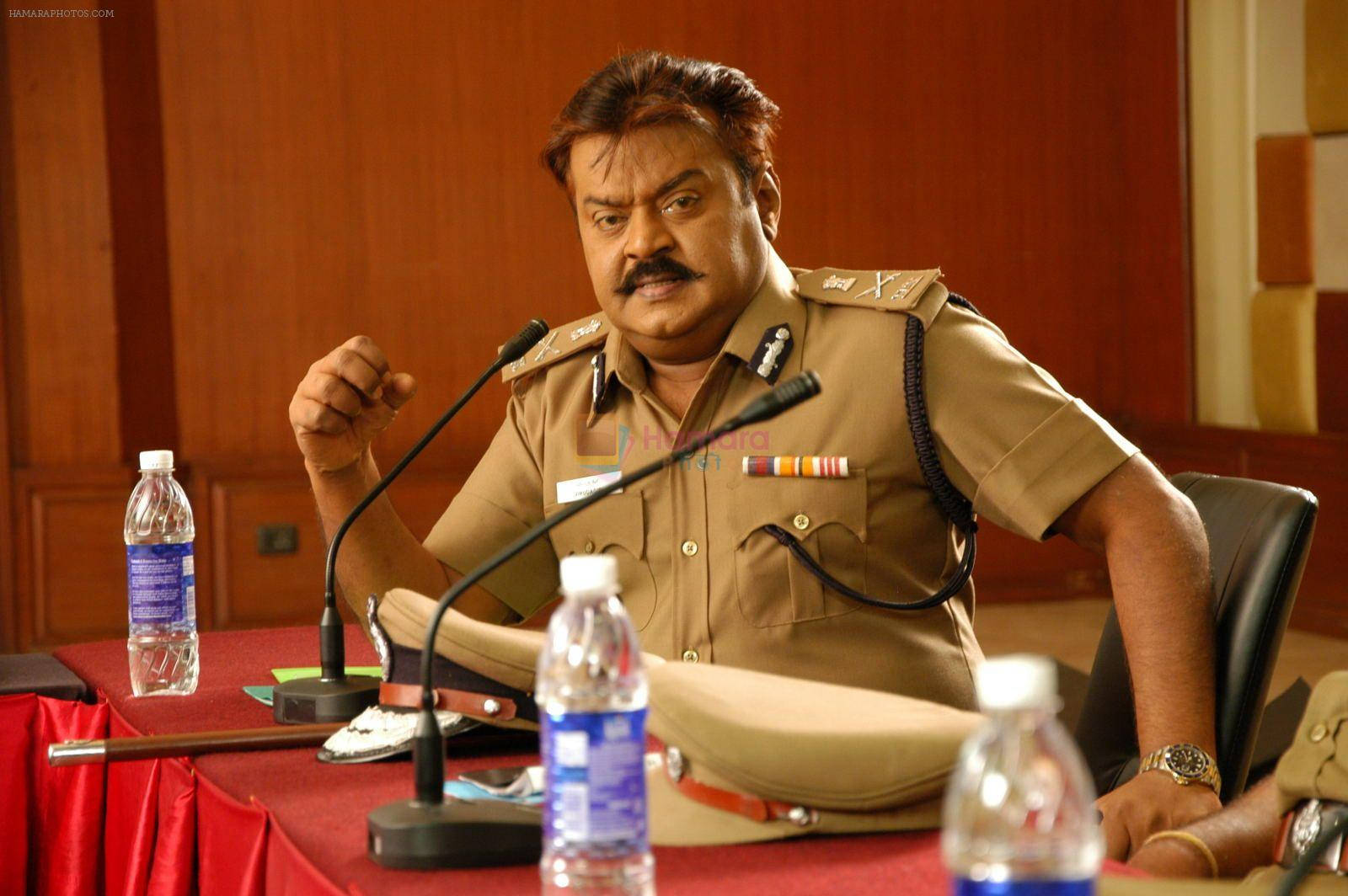 Indian 桌布 壁紙 — 一名身穿卡其色制服、表情嚴肅的警察在記者會上用力做手勢，桌上放著紅色桌布、麥克風和水瓶。