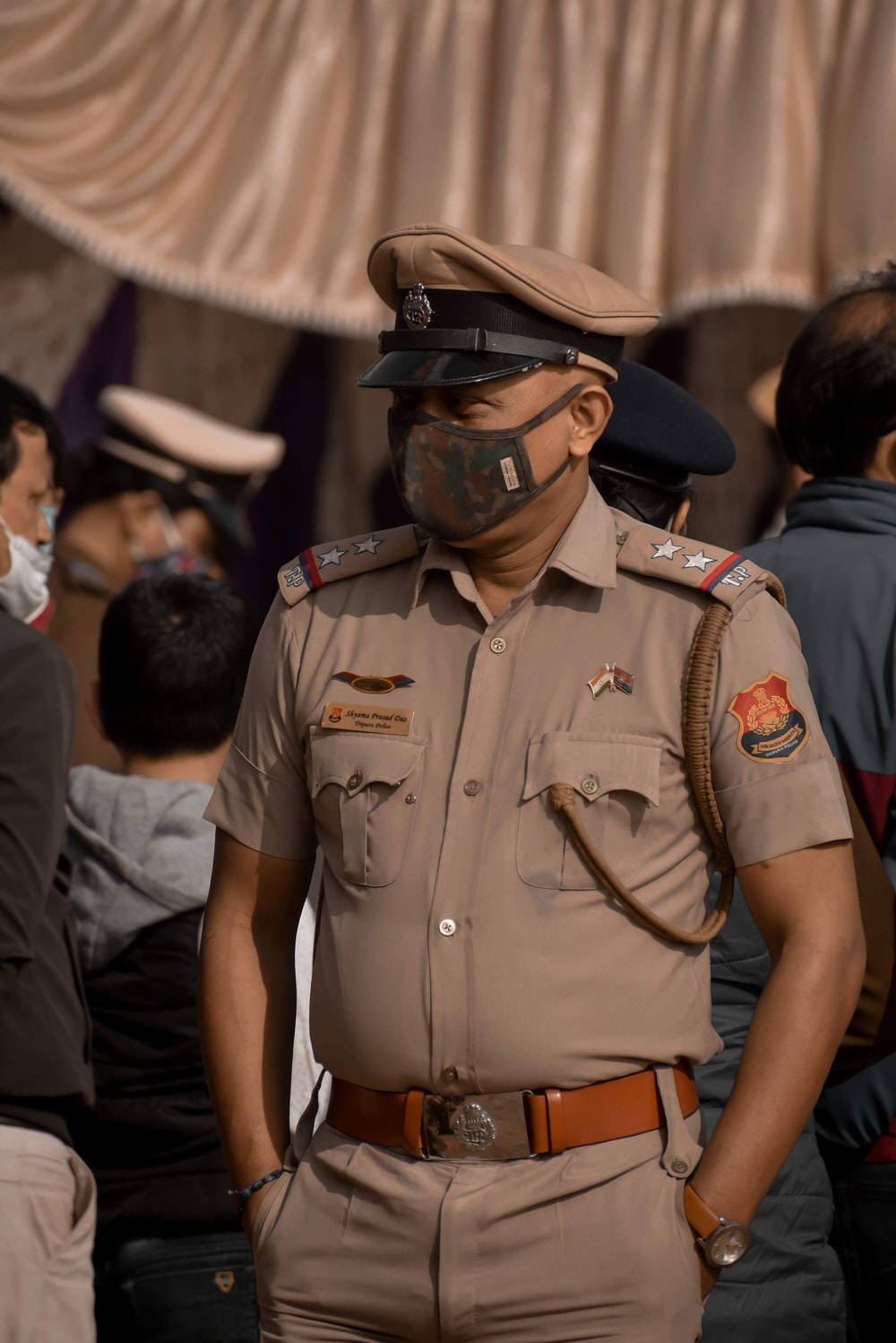 Indian 桌布 壁紙 — 一名身穿卡其色制服及口罩的警察在人群中的布料下保持警覺。