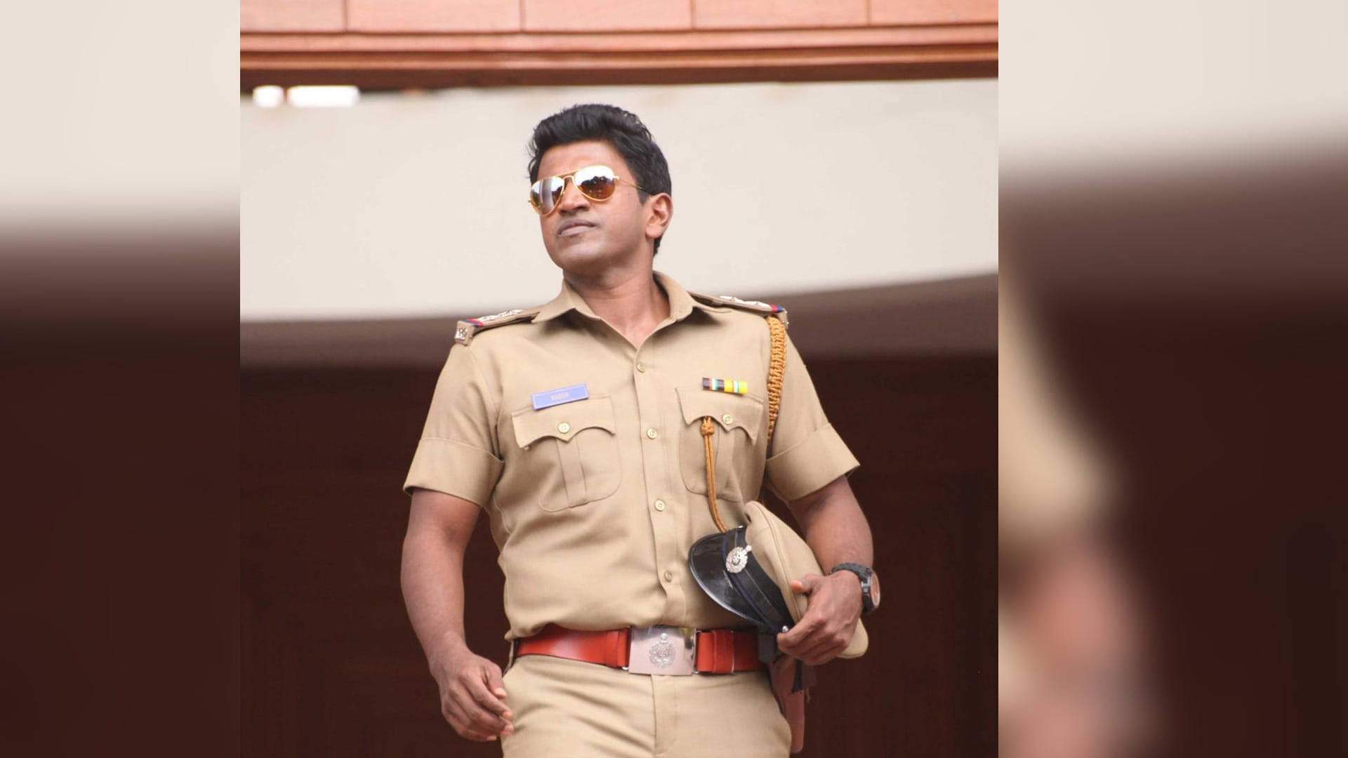 Puneeth Rajkumar 桌布 壁紙 — 一名身穿卡其色制服和太陽眼鏡的警察自信地站立，手持帽子，背景是模糊的建築物。
