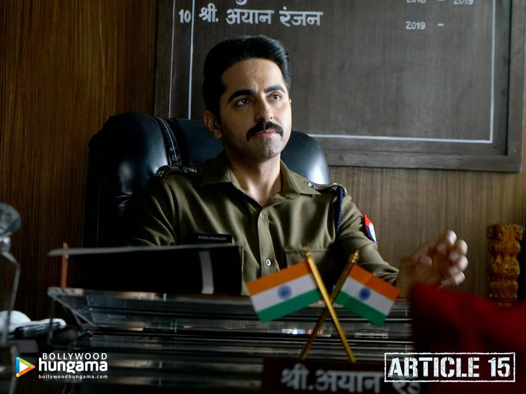 ayushmann khurrana 桌布 壁紙 — 一名身穿卡其色制服的男人坐在玻璃桌前，桌上有印度國旗和名牌，在一個木質牆板辦公室裡。